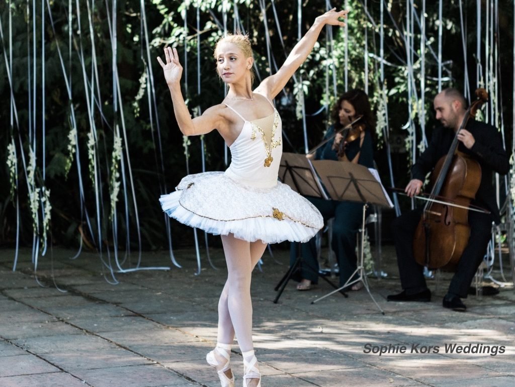 Bailarina de danza clásica durante la recepción de un cóctel de boda, acompañada por músicos de cuerda en directo, creando una atmósfera elegante y delicada.