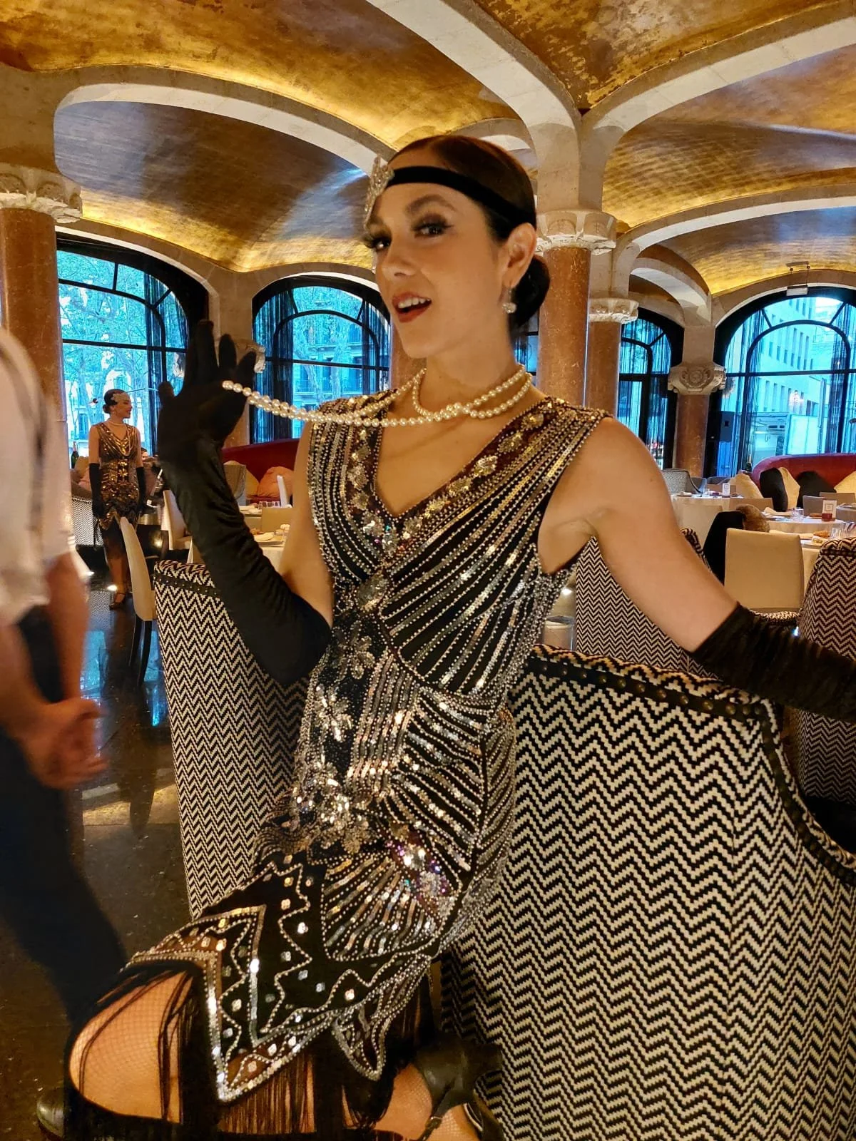 Mujer vestida con estilo de los años 1920, con vestido de flecos y brillo, lentes de tocado de rubí, guantes largos negros y perlas, en un restaurante elegante, en un estilo de época