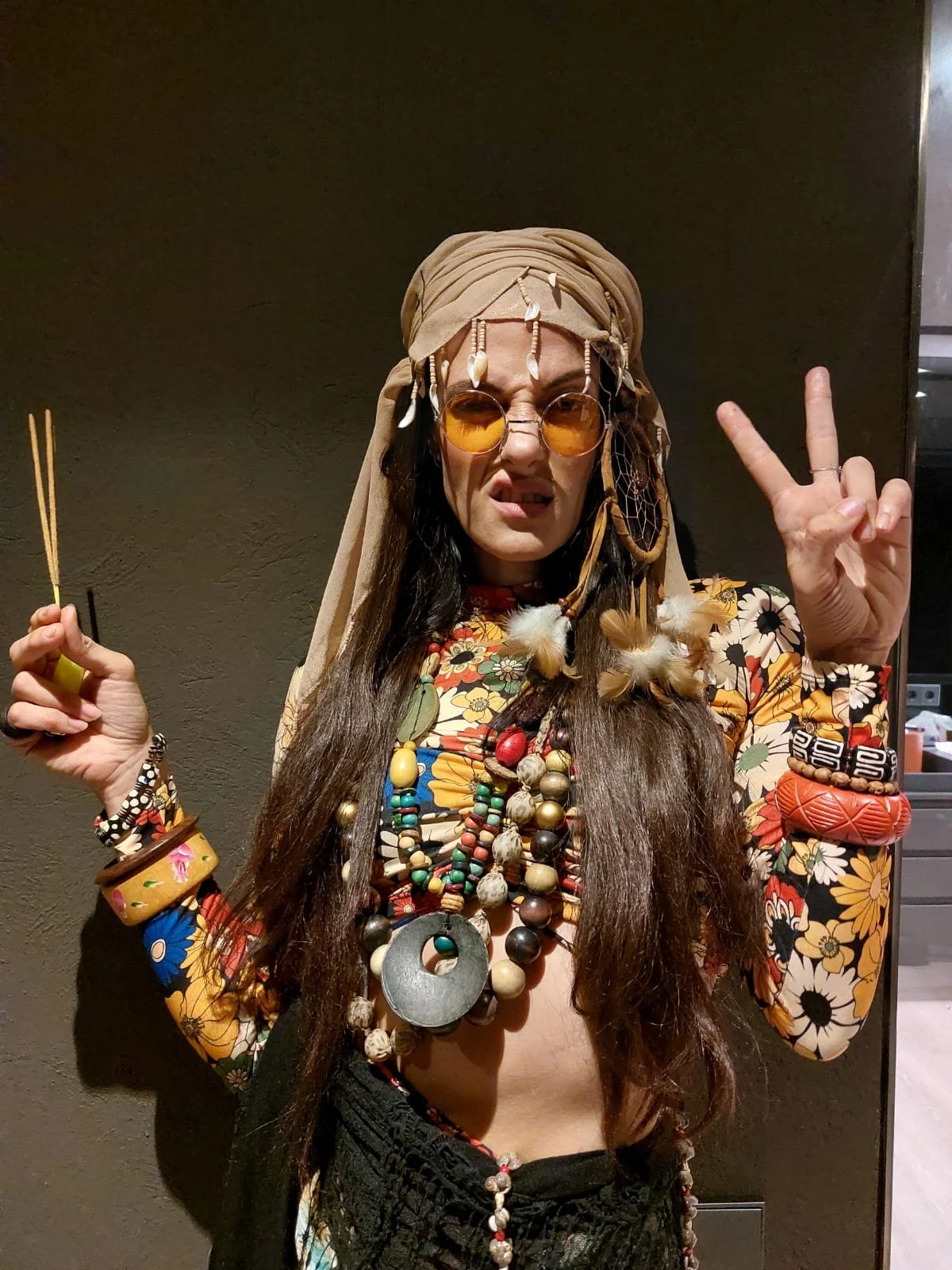 Persona disfrazada con accesorios tribales, gafas de sol anaranjadas, y ropa colorida con estampados florales, haciendo una señal de paz y sosteniendo una varita.