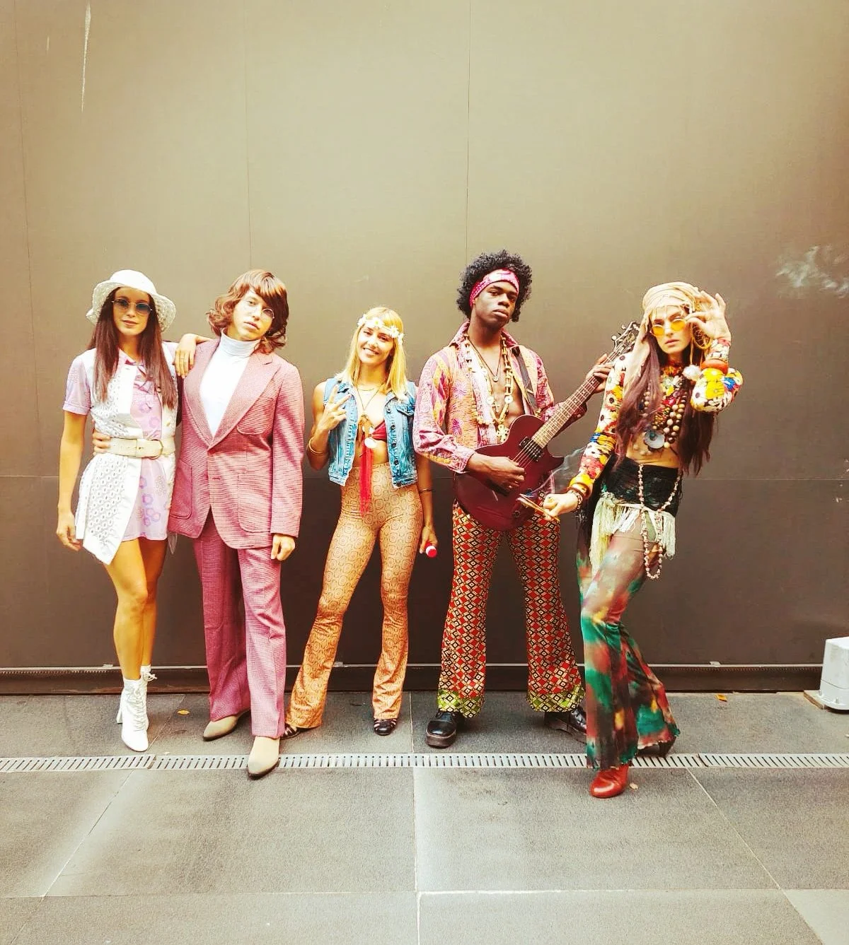 Grupo de cinco personas con atuendos coloridos y estilo bohemio, algunos con instrumentos musicales, de pie contra un fondo simple.