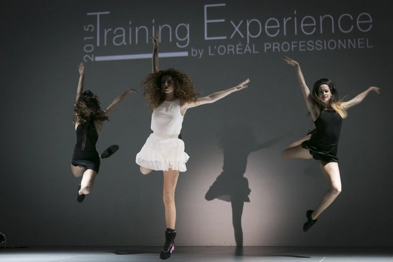 Tres bailarinas de Cuatr8chos saltando en una zona de escenario con fondo de proyección que dice '2015 Training Experience by L'Oréal Professionnel'.