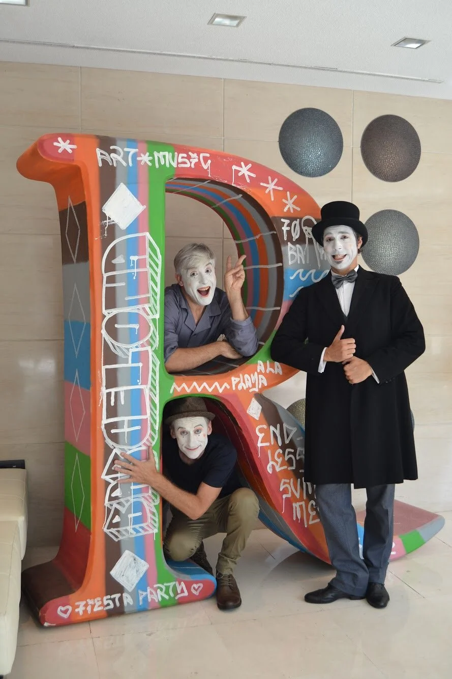 Tres Mimos con maquillaje y disfraces divertidos, uno con un tubo gigante decorado con colores y mensajes, en un lugar con paredes claras y decoraciones de bolas negras.