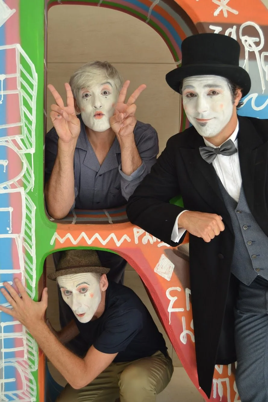 Tres MImos con rostros pintados de blanco y ropa colorida, uno con sombrero de copa, uno con cabello rubio y otro con sombrero de ala ancha. Están en un espacio decorado con colores y frases.