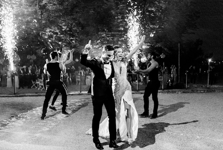Una pareja bailando en una celebración nocturna, rodeados de amigos, con fuegos artificiales en el fondo.