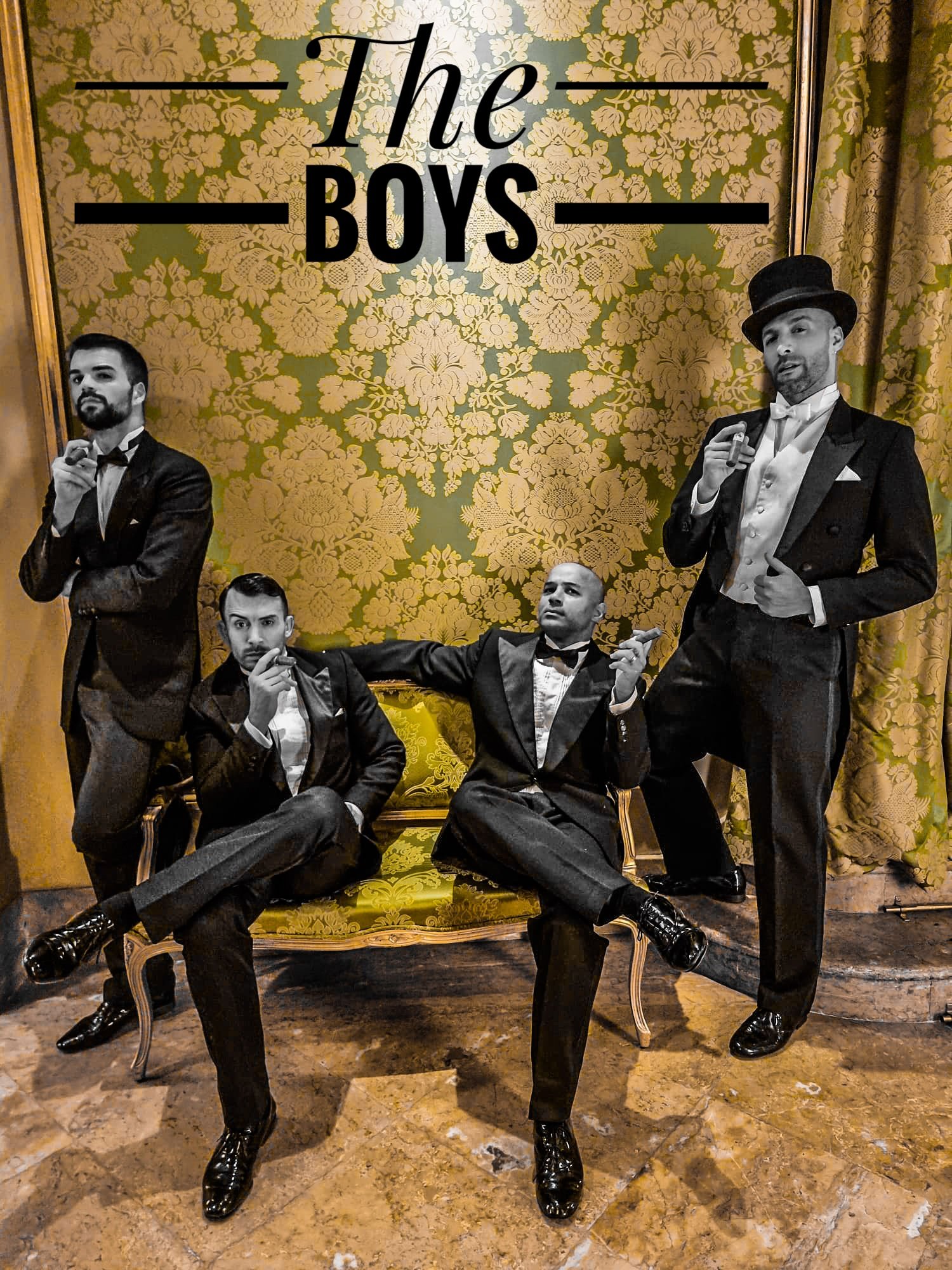 Cuatro hombres vestidos con trajes elegantes y sombreros de copa sosteniendo habanos, posando frente a un fondo con cortinas doradas y papel tapiz con diseño floral, con un cartel que dice "The Boys".