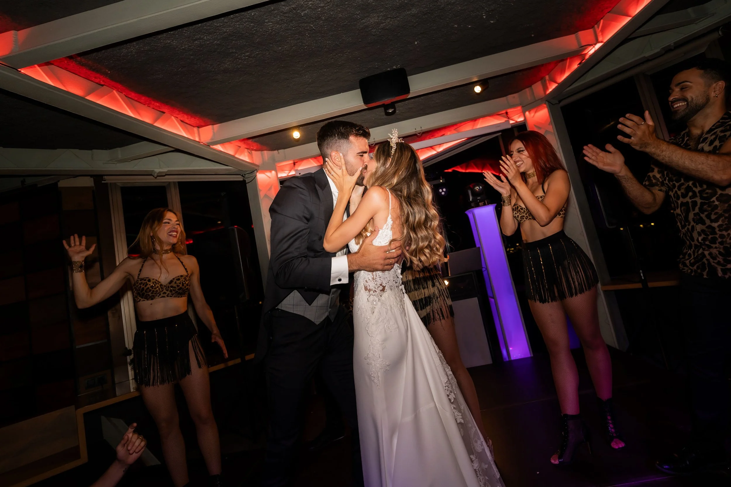 Pareja de novios besándose en su boda, rodeados de amigos que aplauden en un ambiente festivo con iluminación roja y música.