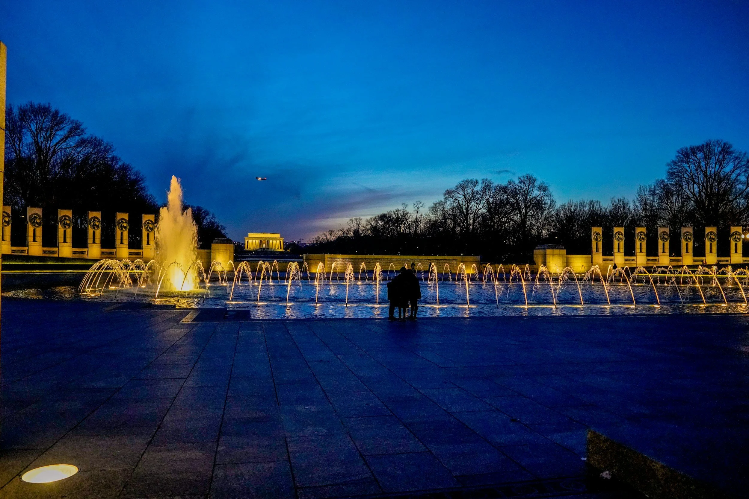 The WWII Memorial in DC at night time, the fountain is running, people are gathered, and the lights are illuminated.