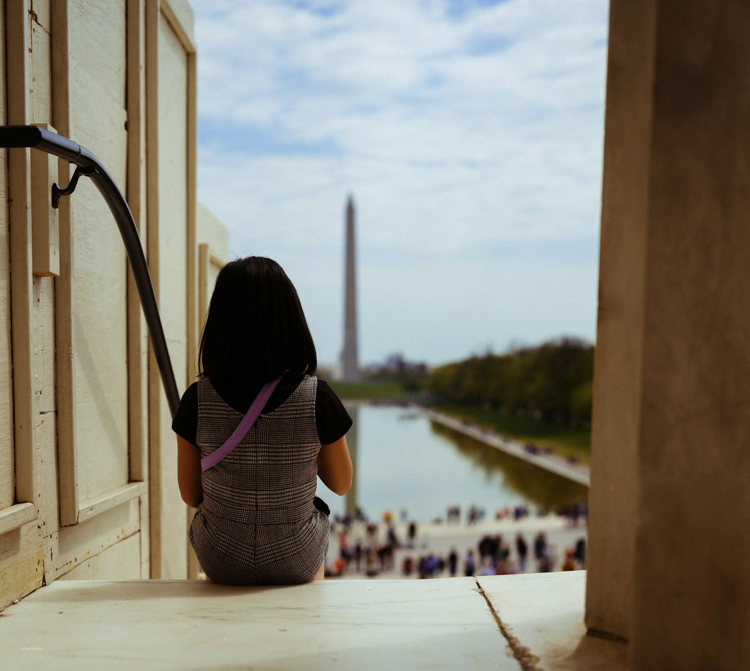 A young girl is sitting on the steps of the Lincoln Memorial in DC and looking towards the Reflection Pool and Washington Monument.