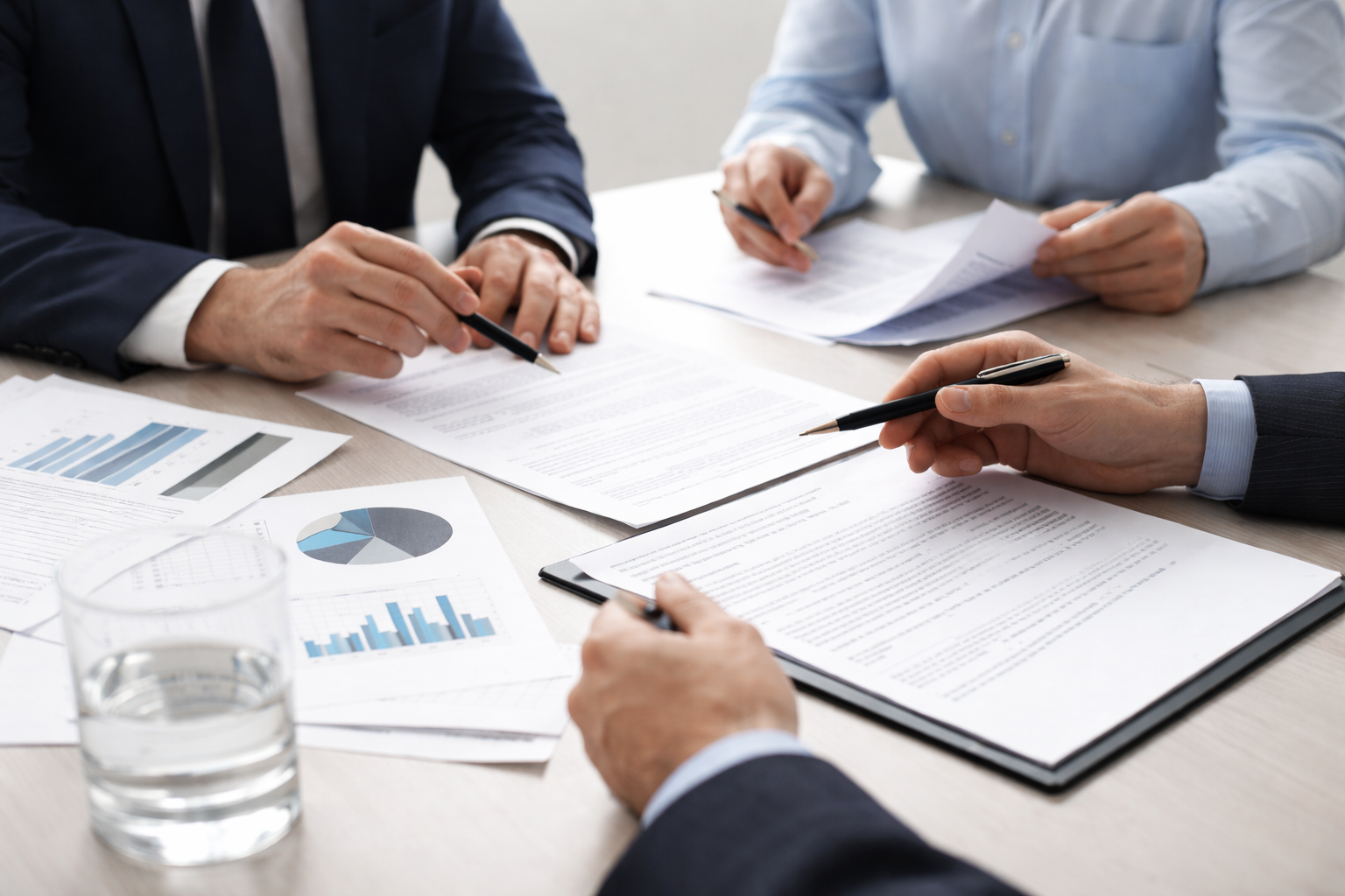 Business professionals sitting at a table reviewing documents and charts, with pens in hand, in a meeting or discussion.