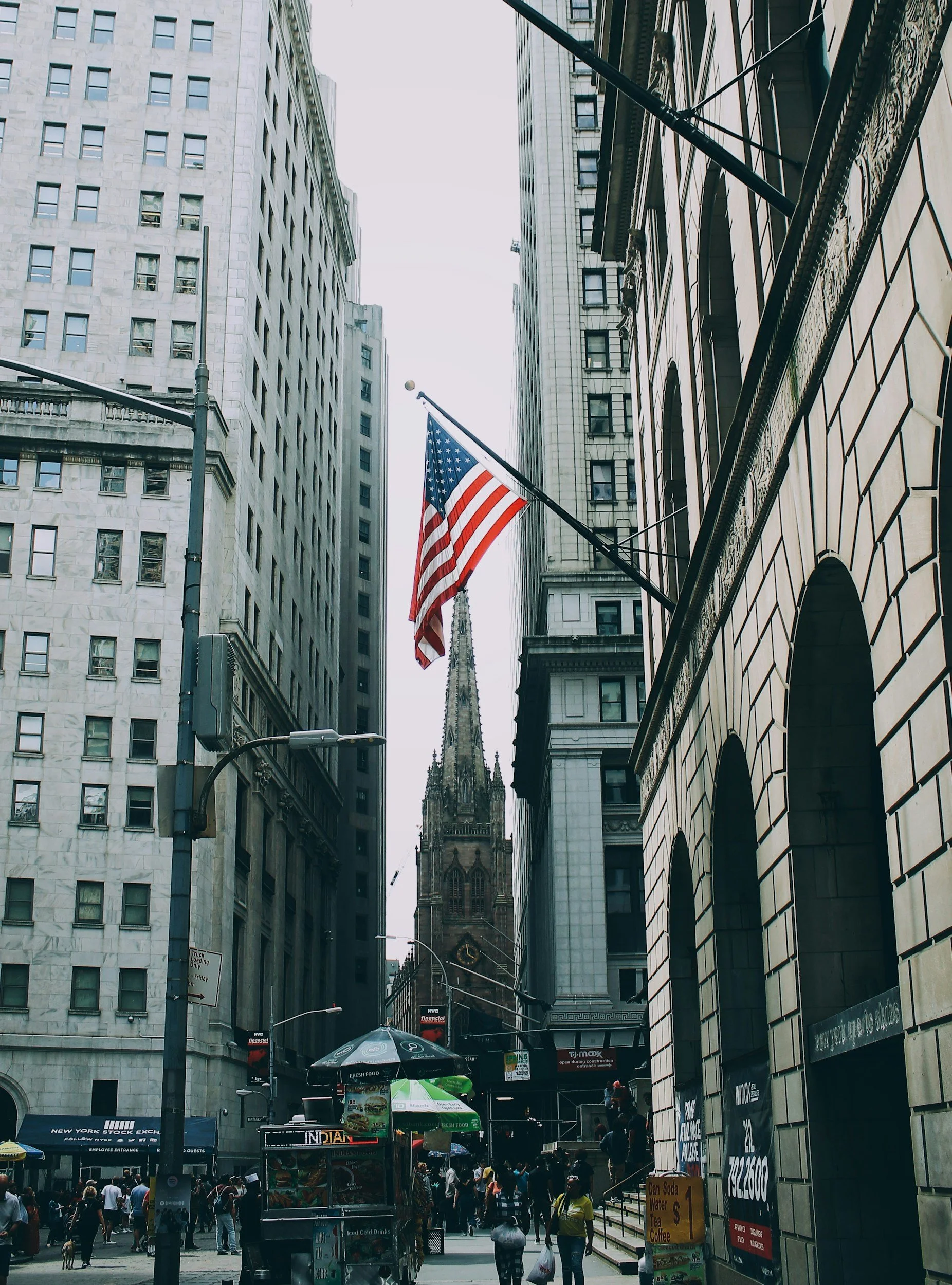 Tall buildings surround a busy street with pedestrians. An American flag flies in the foreground, and a church steeple is visible in the background.