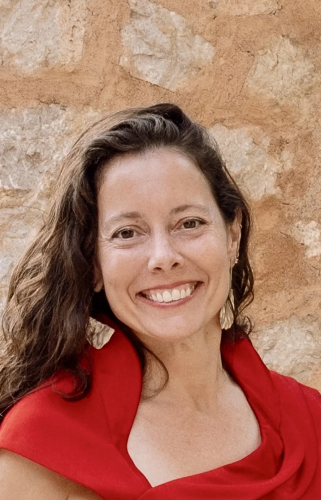 Portrait of a smiling woman with long, wavy brown hair wearing a red top and earrings, standing against a textured stone wall.