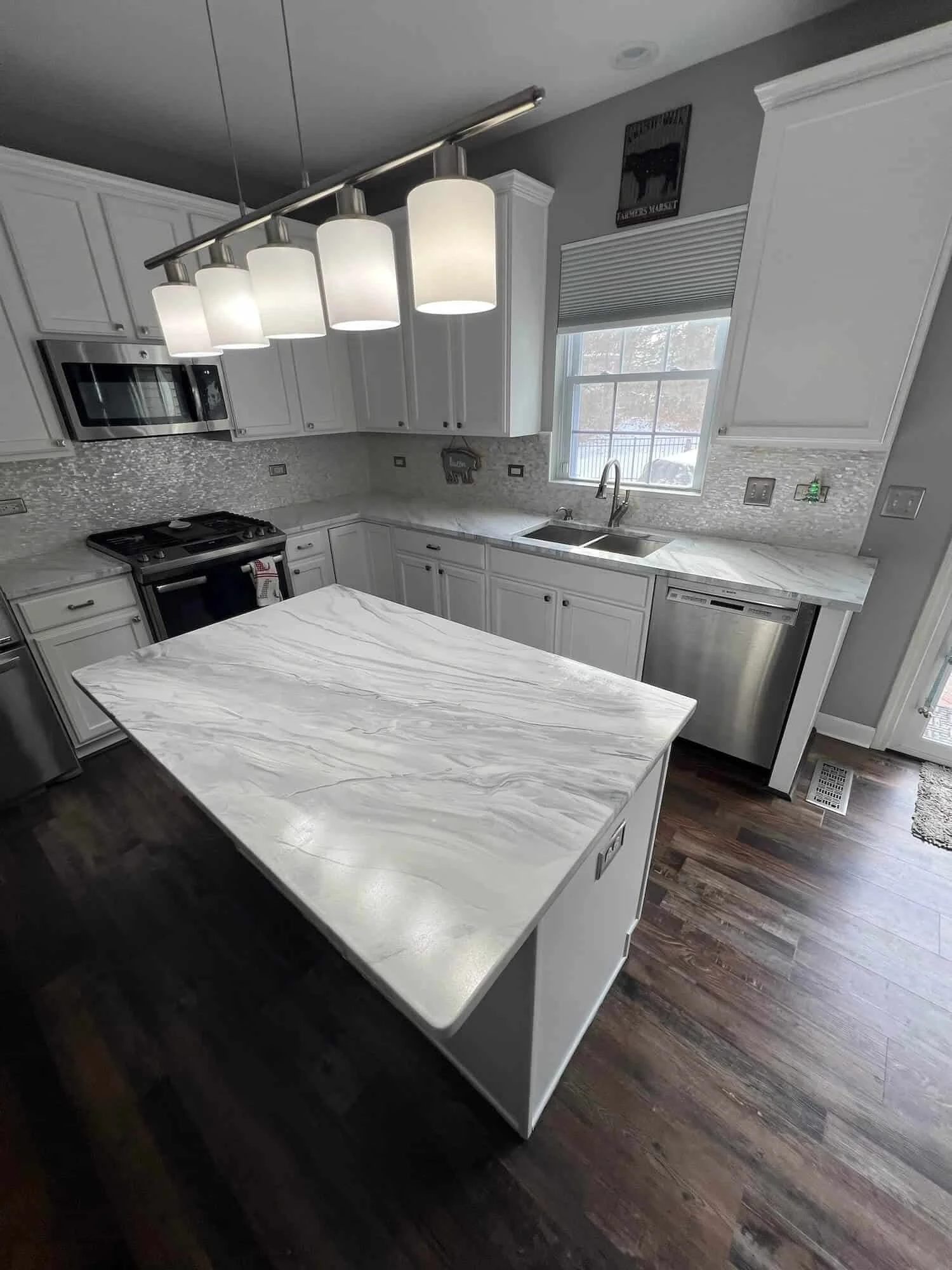 Modern kitchen with white cabinets, marble countertops, stainless steel appliances, dark wooden floor, and a large island with marble surface. Features hanging pendant lights over the island, a window above the sink, and a gray wall with framed decorative artwork.