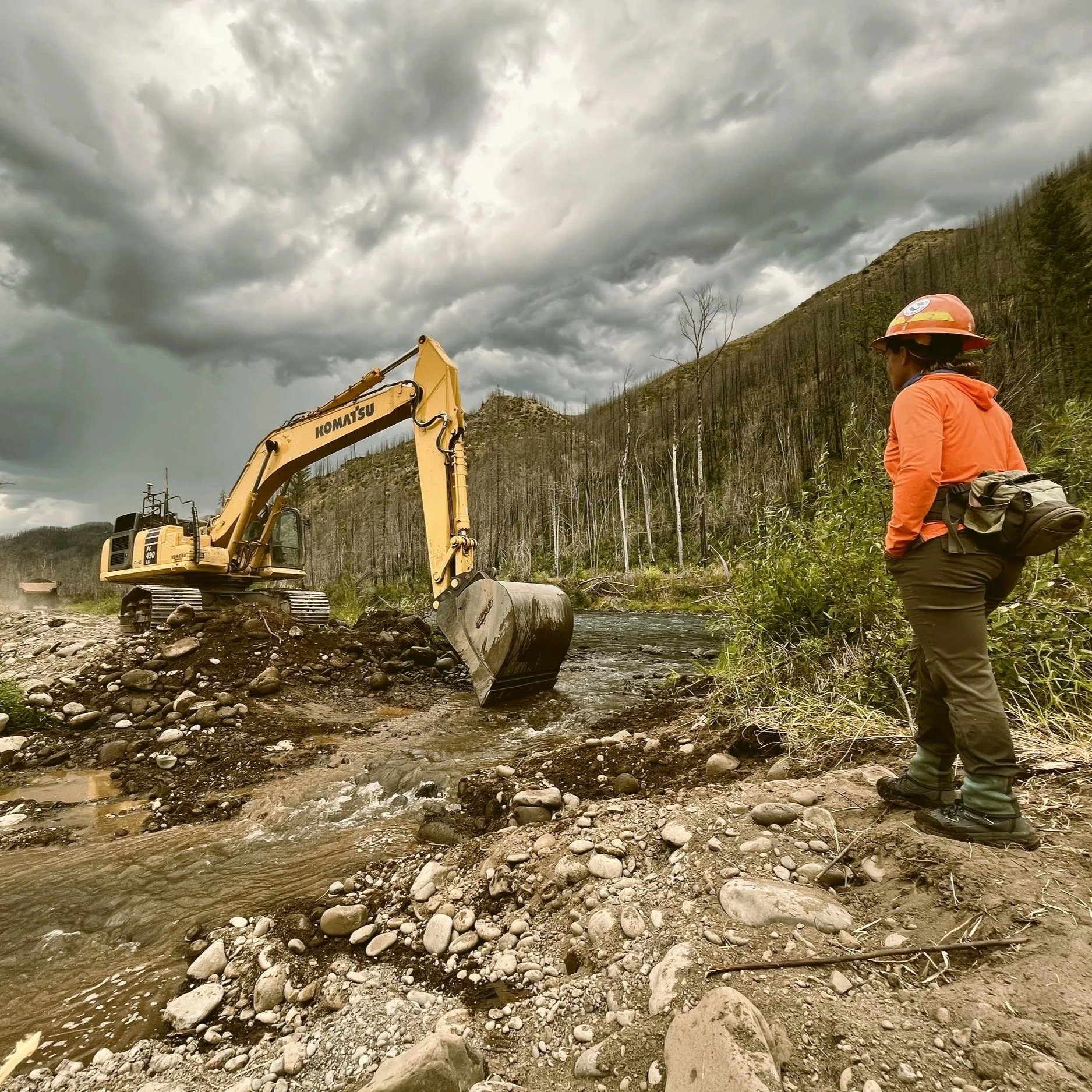 A person wearing an orange jacket and hat stands on a rocky riverbank observing a yellow excavator moving rocks and debris in a river. The scene is set against a backdrop of cloudy skies and a hillside with sparse trees.