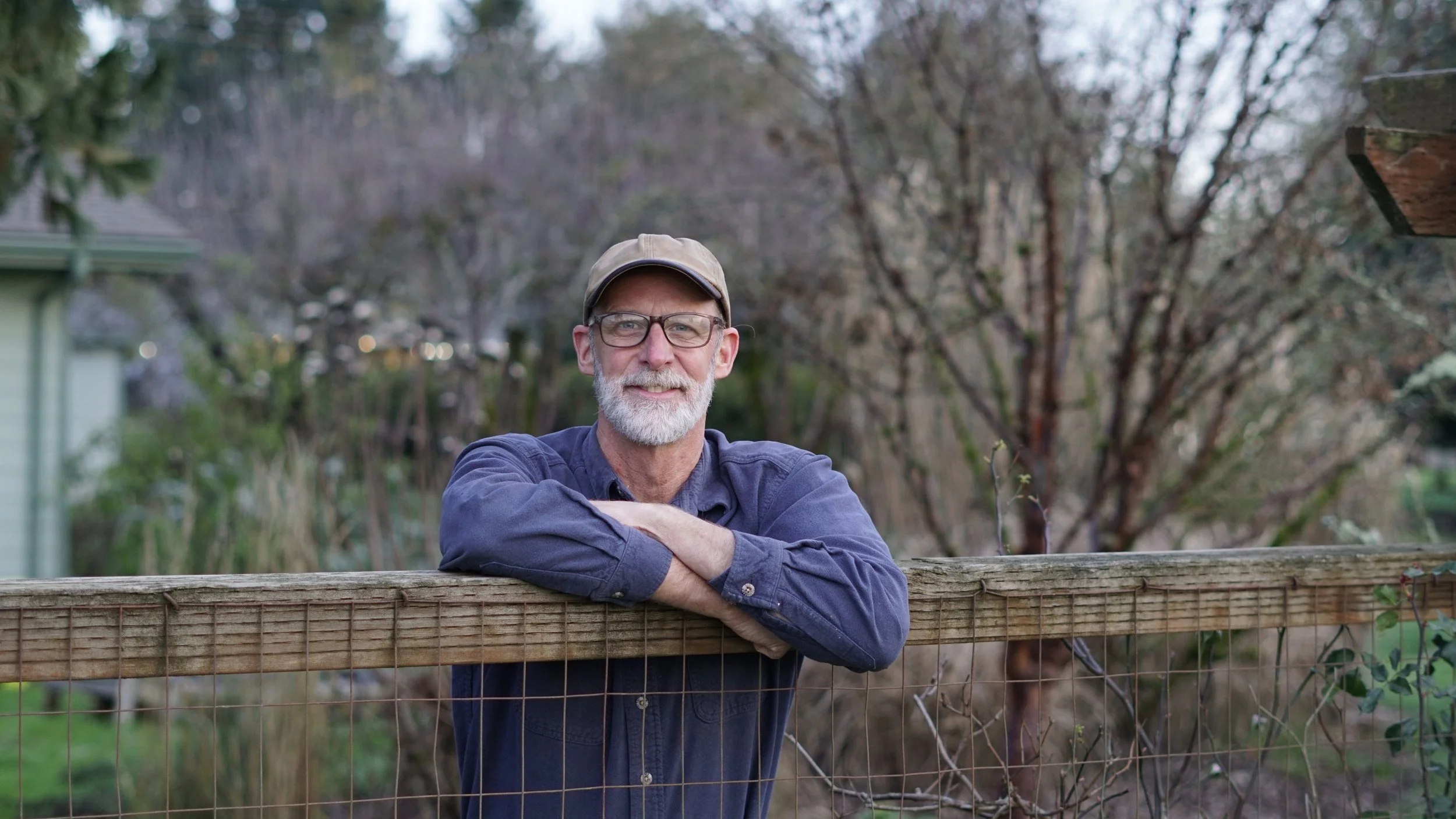 A man with glasses, a beard, and a cap leaning on a wooden fence outdoors, with trees and a house in the background.