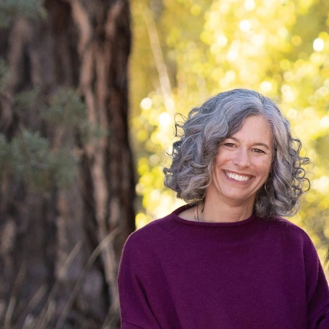 A smiling woman with curly gray hair wearing a maroon sweater outdoors in front of a tree and yellow leafy background.