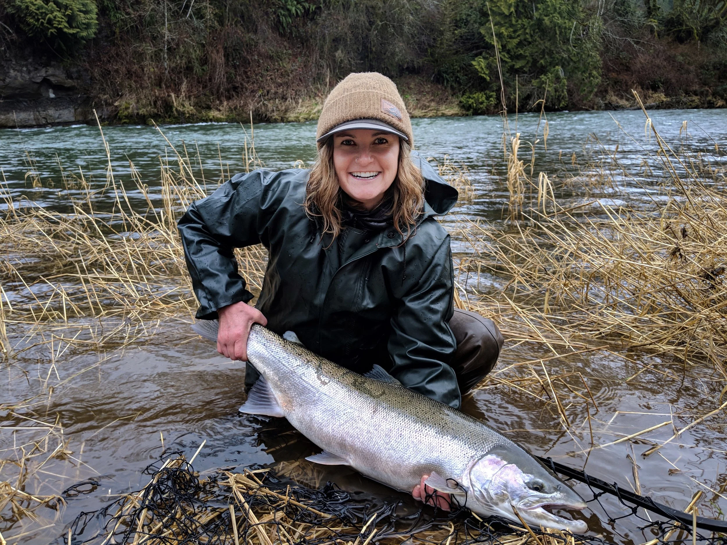 A woman in outdoor gear kneeling in water, holding a large fish she caught while fishing in a river.