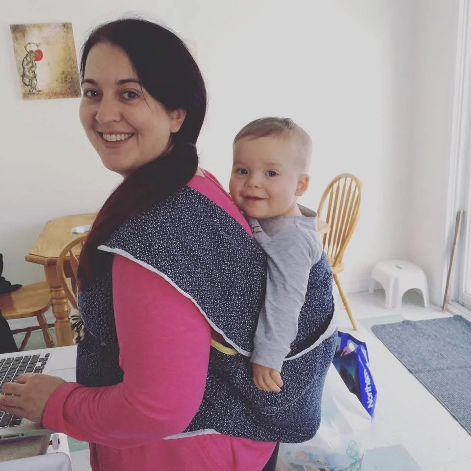 A woman smiling with a baby on her back in a baby carrier in a home kitchen.