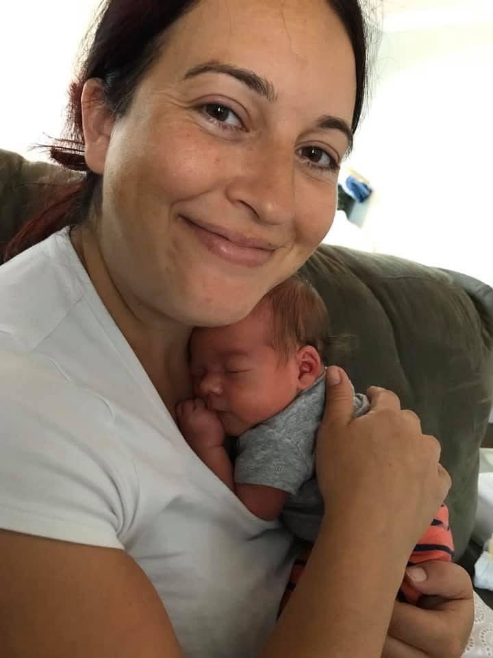 Woman smiling and holding a sleeping baby close to her chest, sitting on a couch in a cozy home setting.