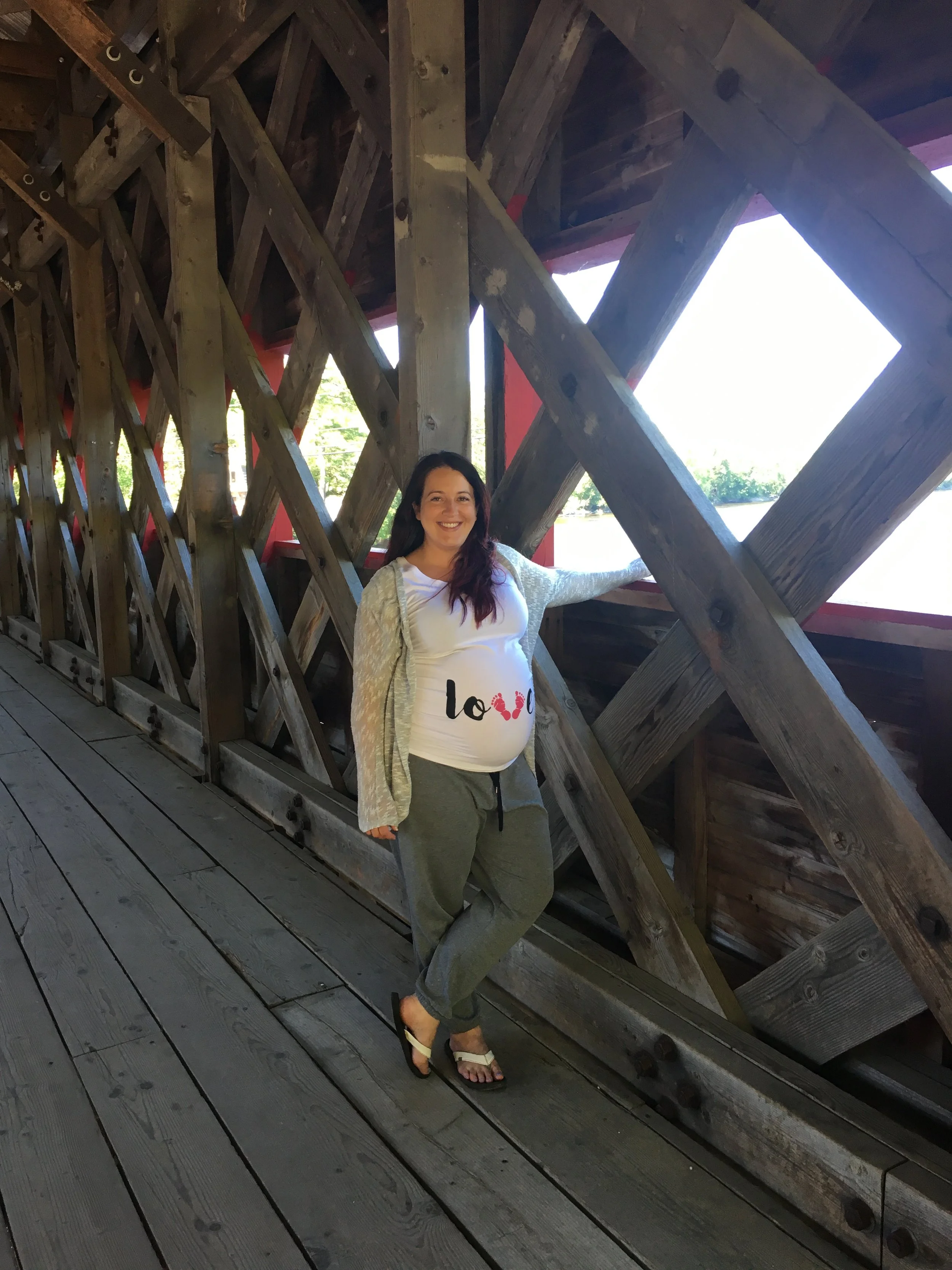 A woman standing inside a wooden bridge with large, crisscrossed beams, smiling and posing near a large square window opening, wearing a white t-shirt with 'love' written on it, gray pants, and flip-flops.