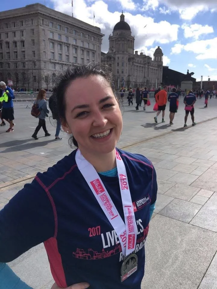 A woman smiling after finishing a race, wearing a medal and a race shirt, in a city square with historic buildings and other runners in the background.