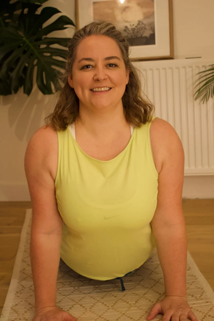 A woman with wavy hair smiling at the camera, wearing a bright yellow tank top, practicing yoga or stretching on a mat indoors with plants and framed artwork in the background.