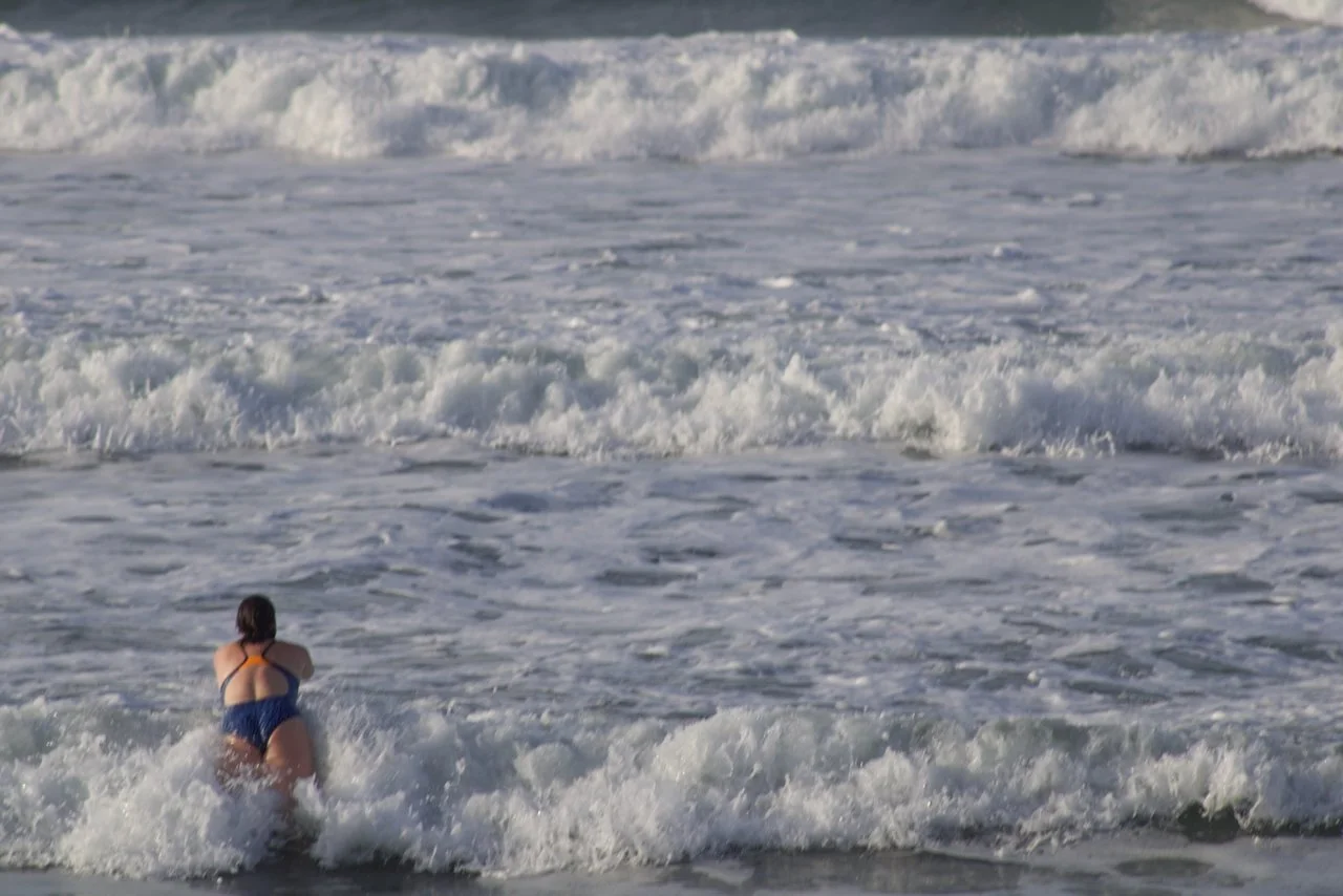A woman in a blue swimsuit is sitting in the ocean waves at the beach.