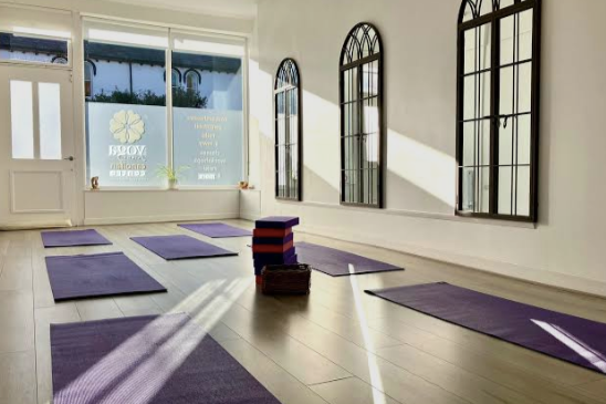 Empty yoga studio with purple mats, a stack of purple yoga blocks, and large arched windows letting in sunlight.