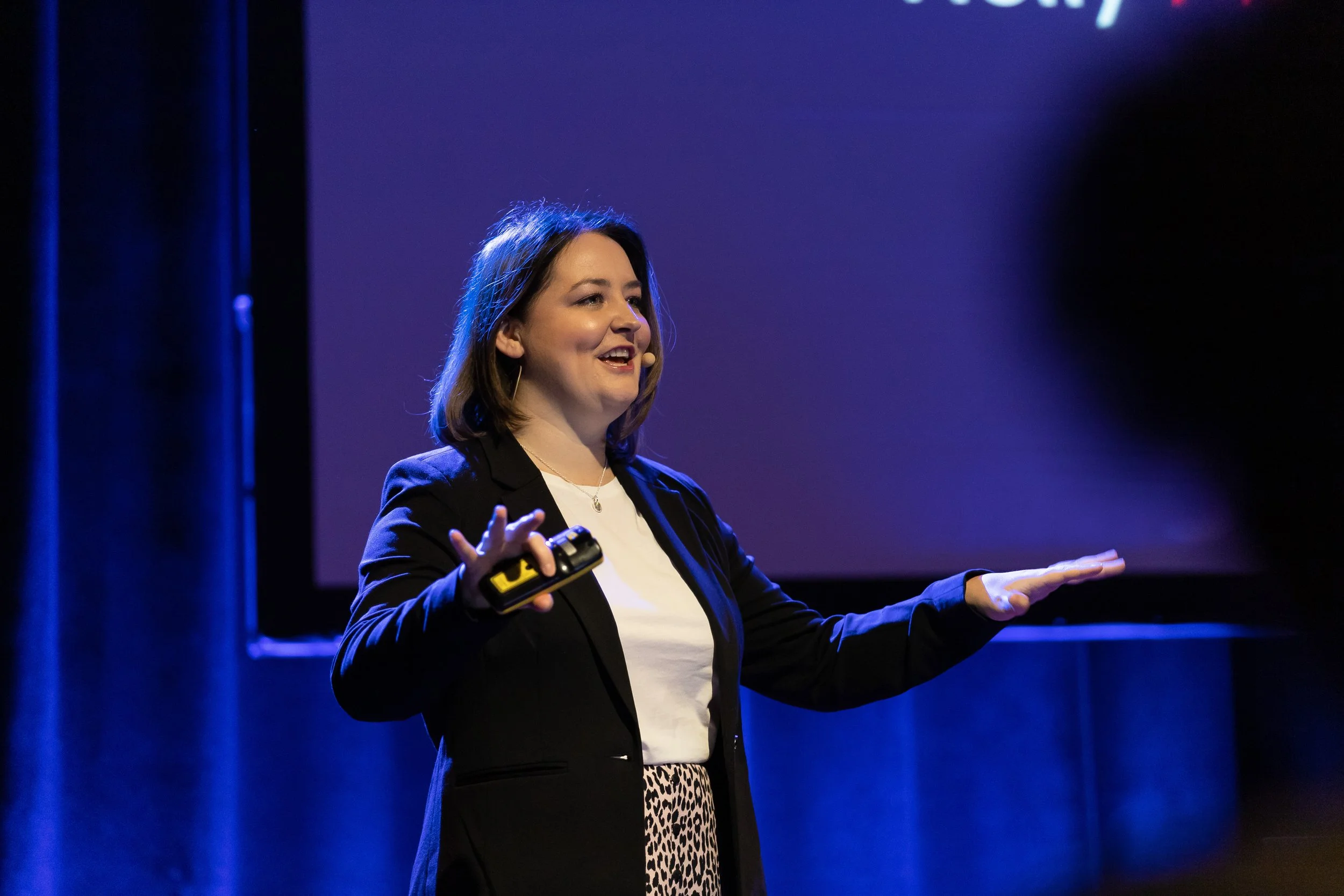 A woman giving a presentation on stage, holding a remote control in her right hand, wearing a black blazer, a white shirt, and a leopard print skirt. The background is dark with purple and blue lighting.
