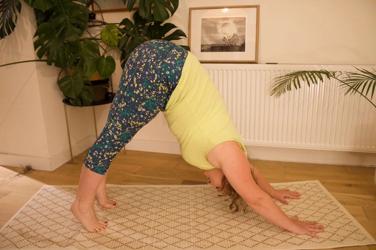 A woman practicing yoga in downward dog pose indoors, on a patterned rug, with potted plants and framed artwork on the wall behind her.