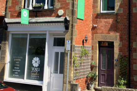 Exterior of a building with a yoga studio sign, showing a green sign with a white lotus and the words 'Yoga Canolfan Centre', and a purple door surrounded by plants.