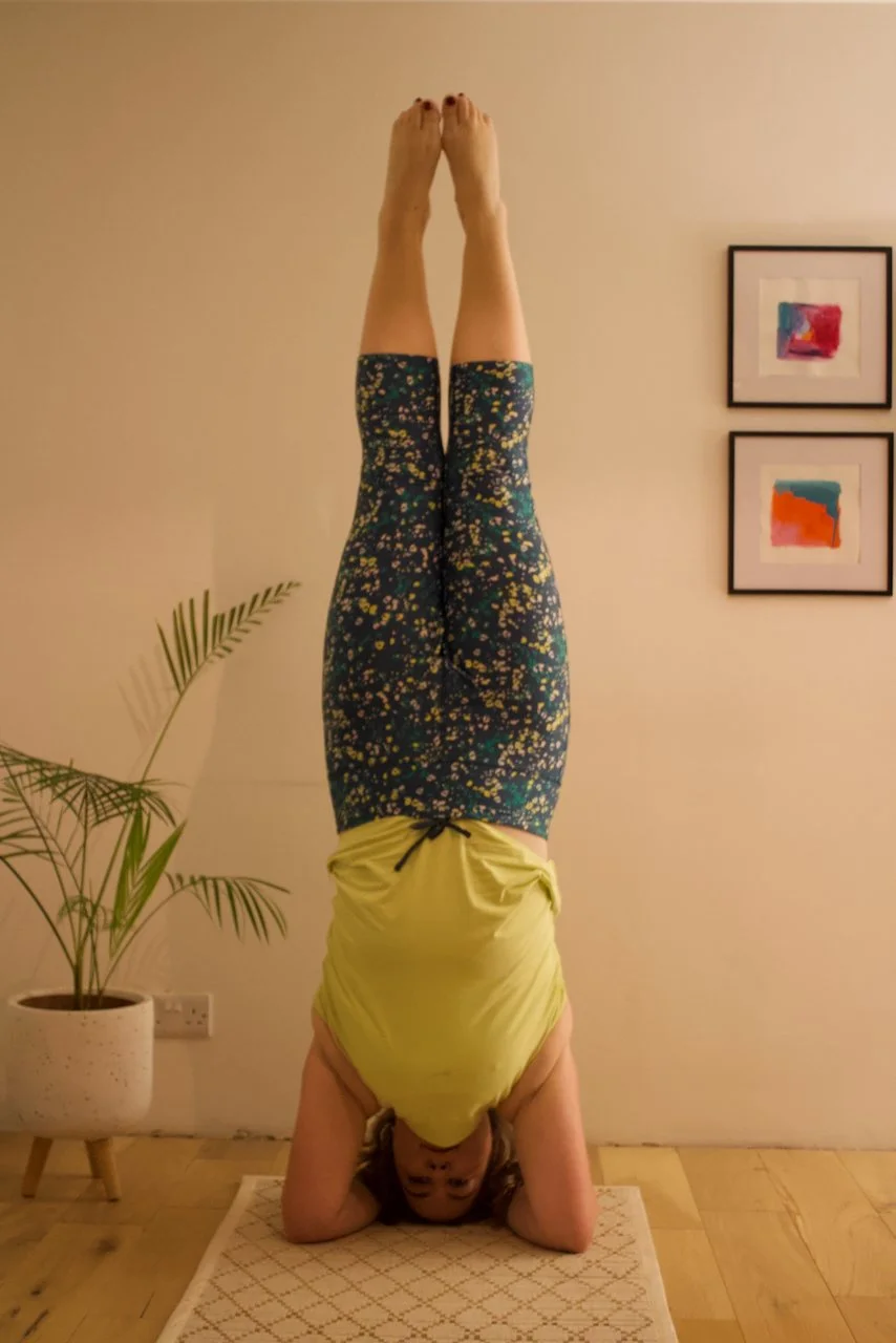 A woman doing a headstand yoga pose indoors on a white patterned rug, wearing a yellow top and dark leggings with colorful splashes. There are framed abstract art pieces on the wall, a potted plant, and a wooden floor.