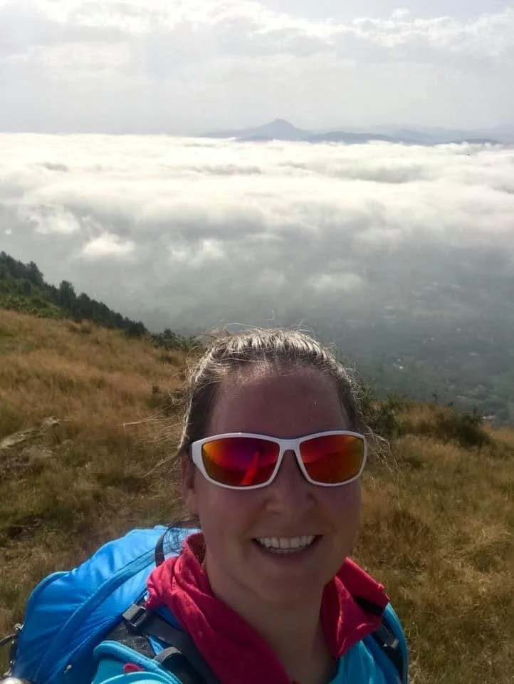 A woman with sunglasses smiling in a mountainous area with grassy terrain, clouds below and a mountain in the distance.