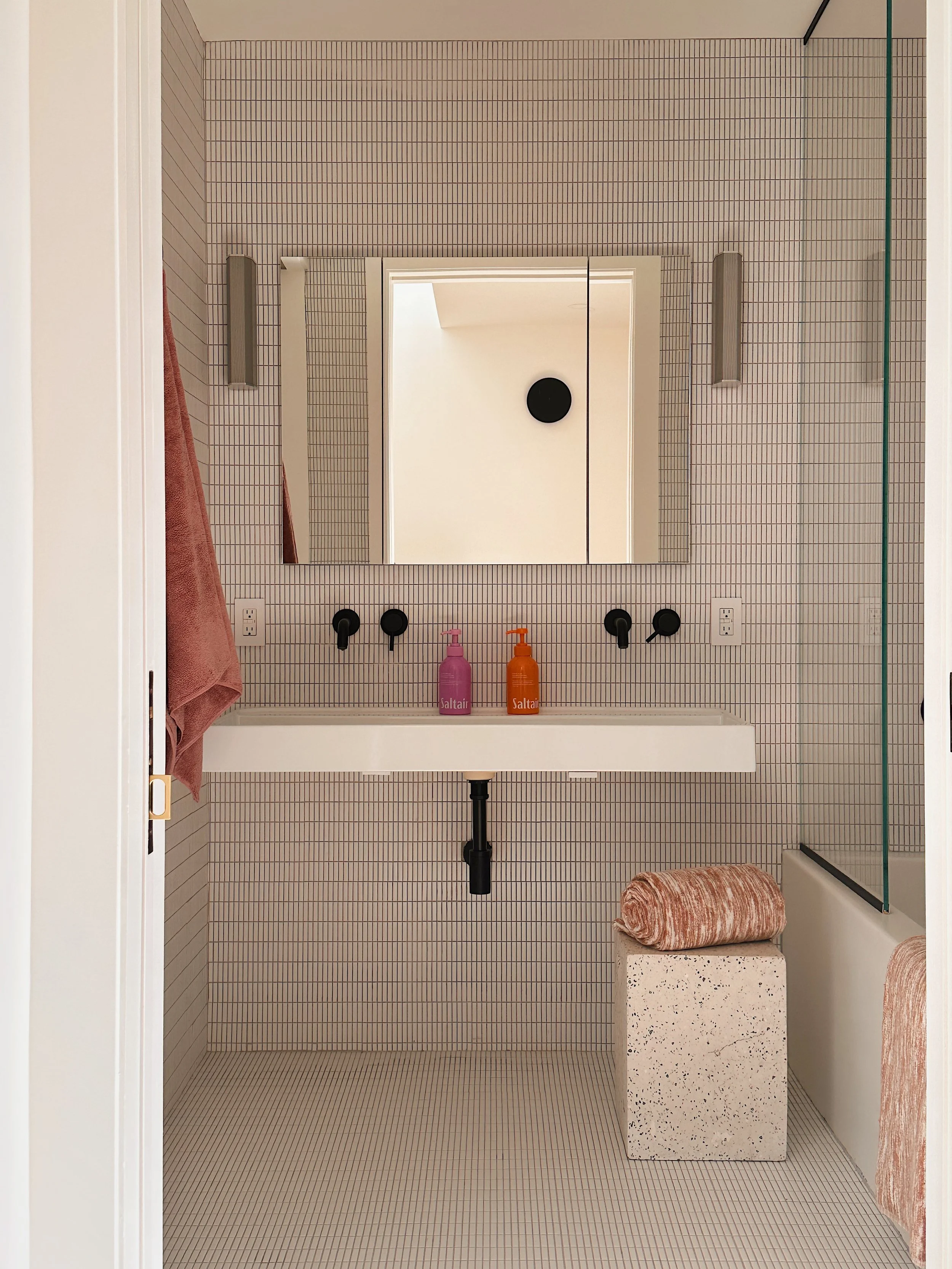 Minimalist bathroom with small square tiles on walls and floor, wall-mounted sink with two bottles of Saltair soap, large mirror, pink towel on the left hooks, ceiling light, and a terrazzo stool with a rolled peach-colored towel beside a bathtub with a glass partition.