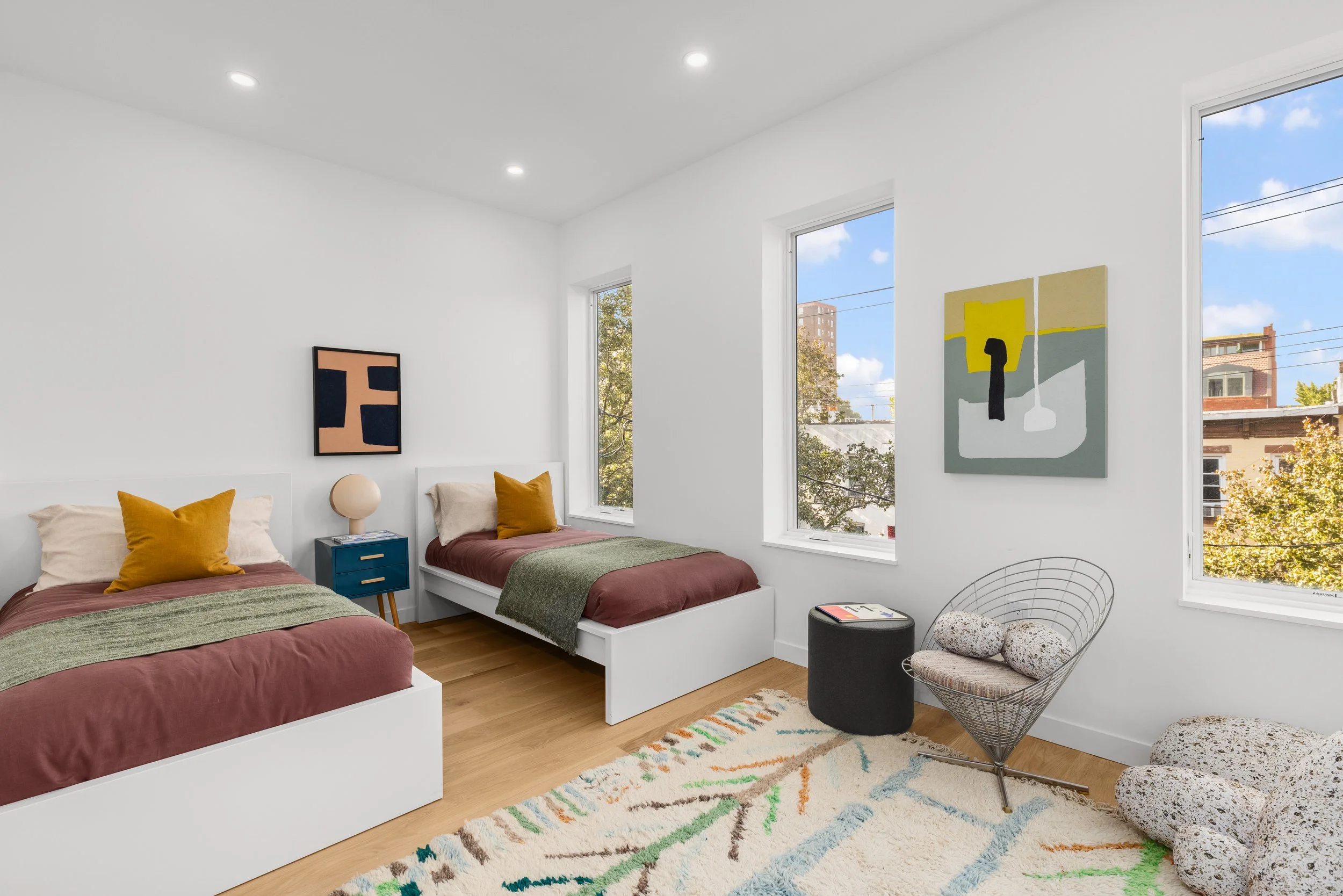 A bedroom with two twin beds, mustard pillows, green blankets, white walls, and large windows showing trees and a building outside. Decor includes abstract art and a modern chair with speckled pillows.