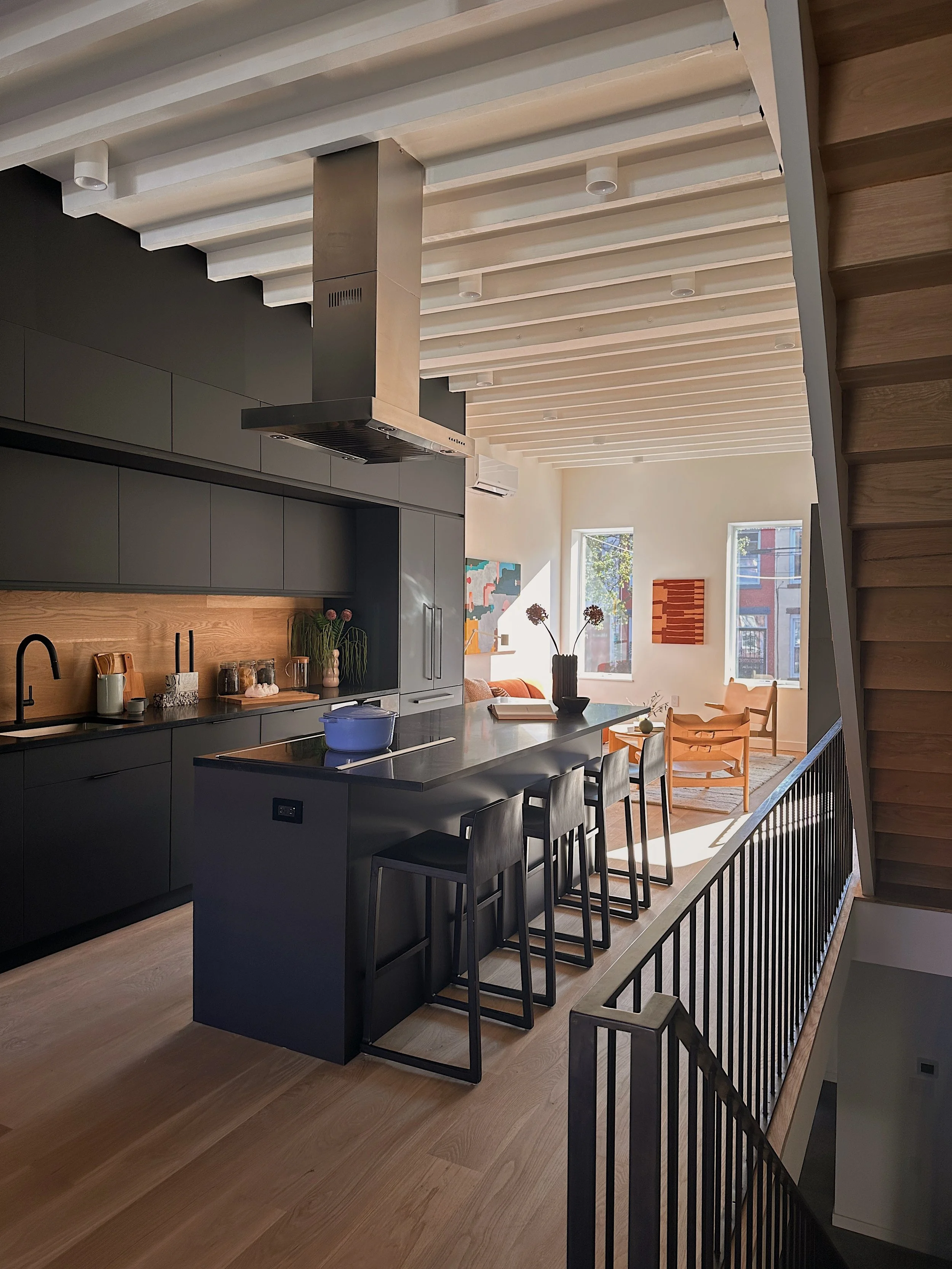 Modern kitchen with dark cabinetry, a large island with black chairs, and a living area with orange chairs and artwork on the wall.