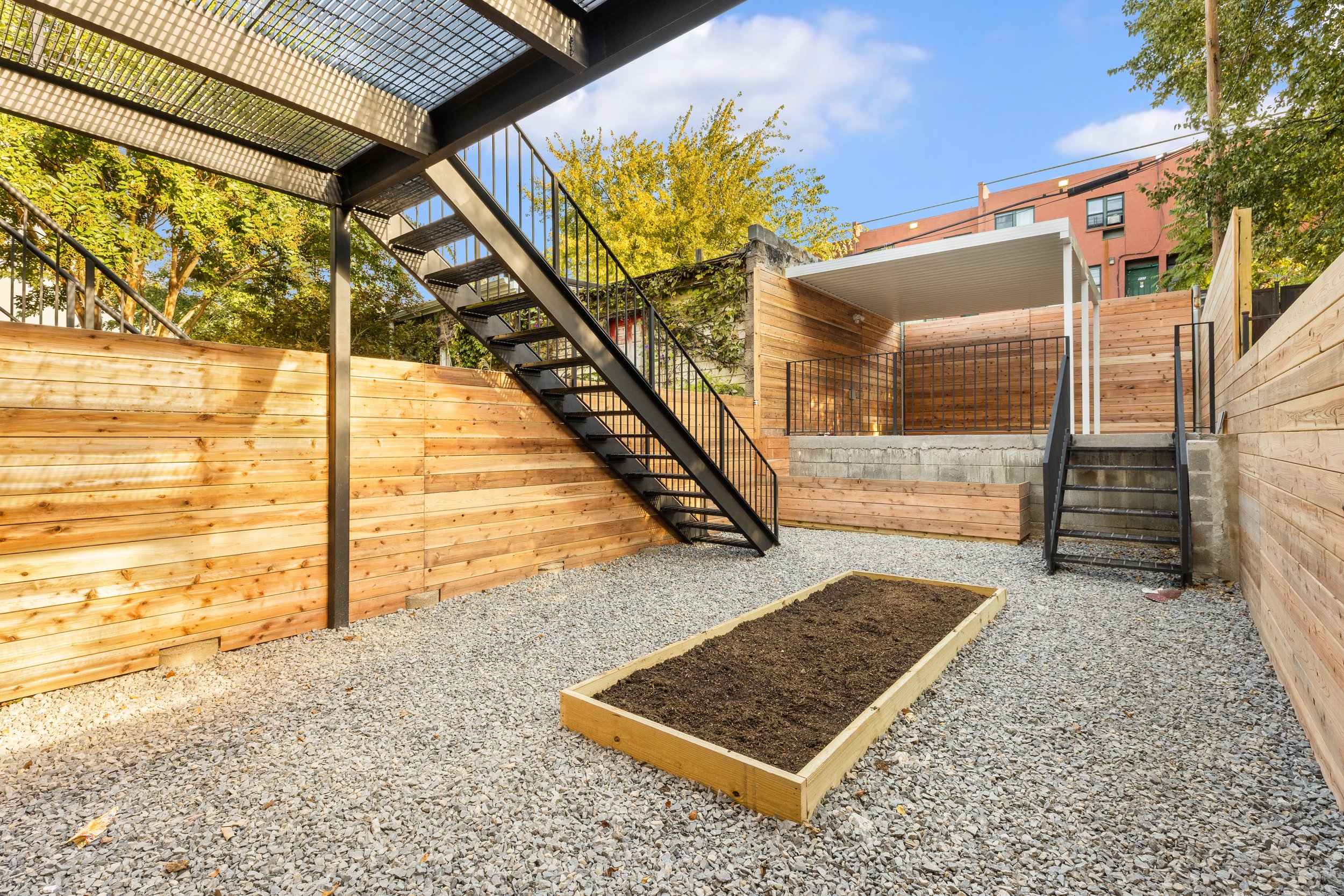 A gravel backyard with a rectangular wooden garden bed filled with soil, surrounded by a wooden fence. There are two staircases, one black metal and one concrete with a black metal railing, leading to an elevated wooden deck area with a white canopy. Trees and a brick building are visible in the background under a partly cloudy sky.