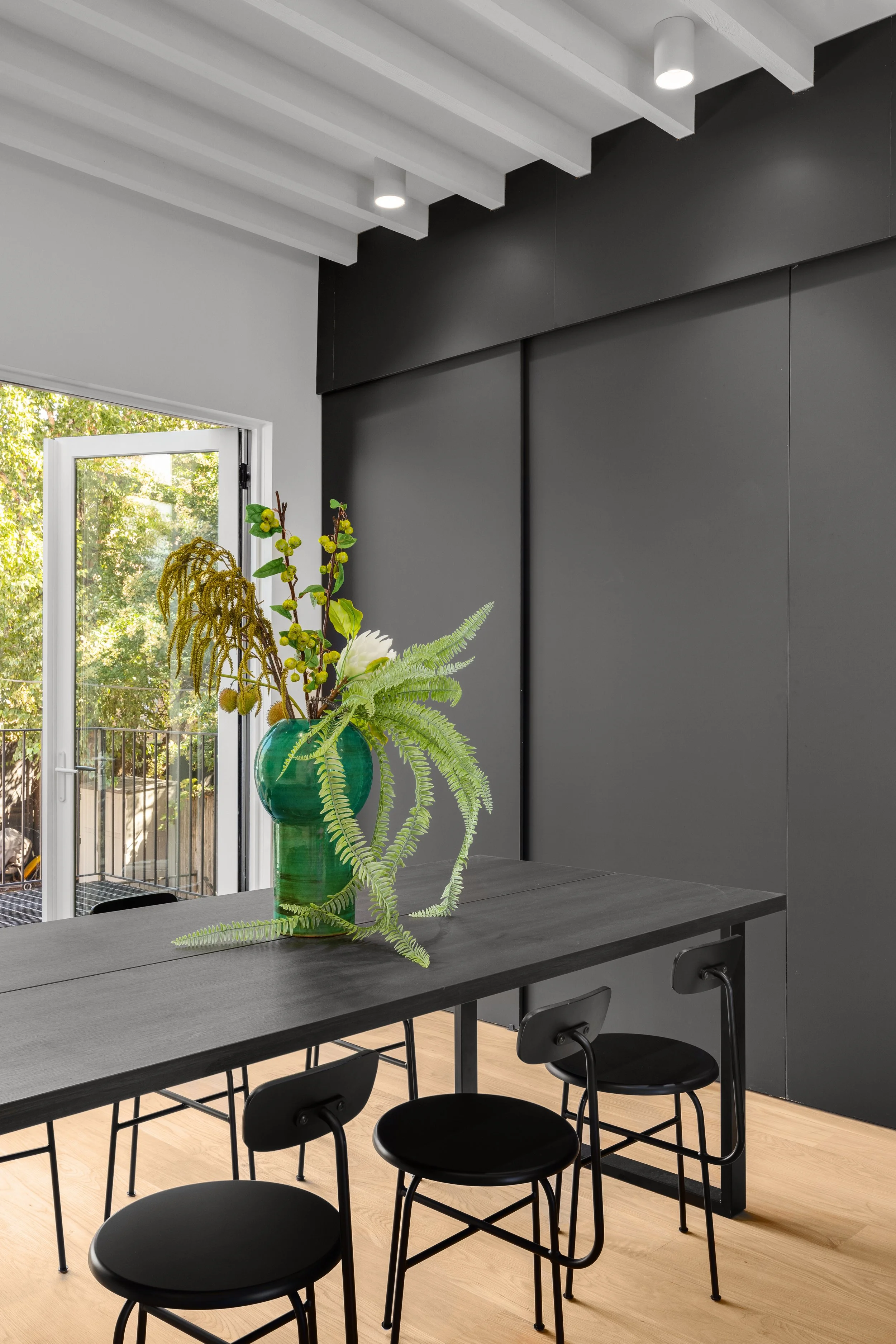 A modern dining area with a black table, four black chairs, and a green vase with various green plants on the table. There's a glass door leading outside, with trees visible outside. The ceiling has white beams and two built-in lights. The wall behind the table is painted dark gray.