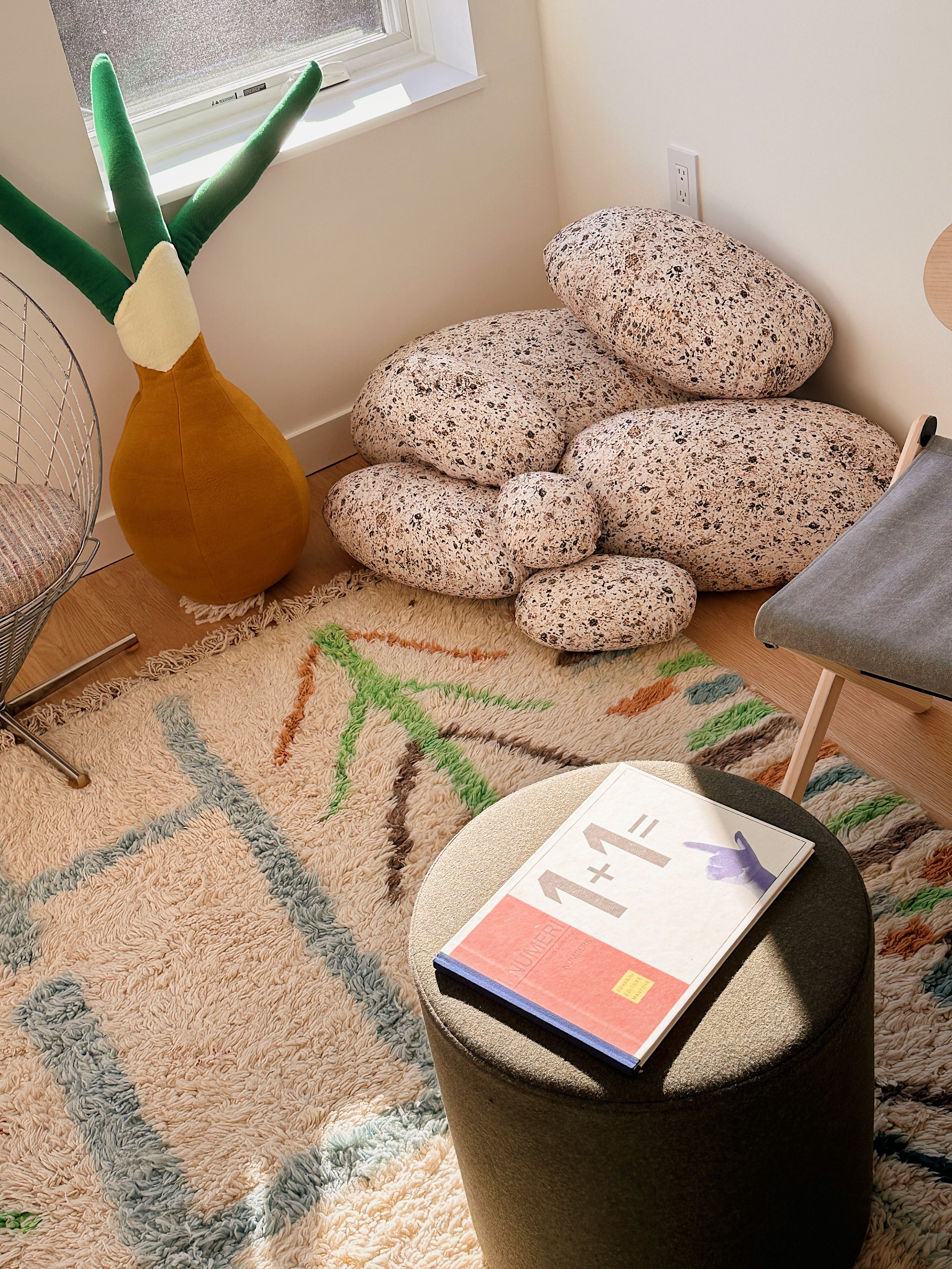 Room corner with a plush, cream-colored rug with a multicolored abstract pattern, a black cylindrical stool with a book on top, a gray chair, a decorative pouf made of large speckled stones, a yellow floor vase with green fabric plants, and a window with white trim.