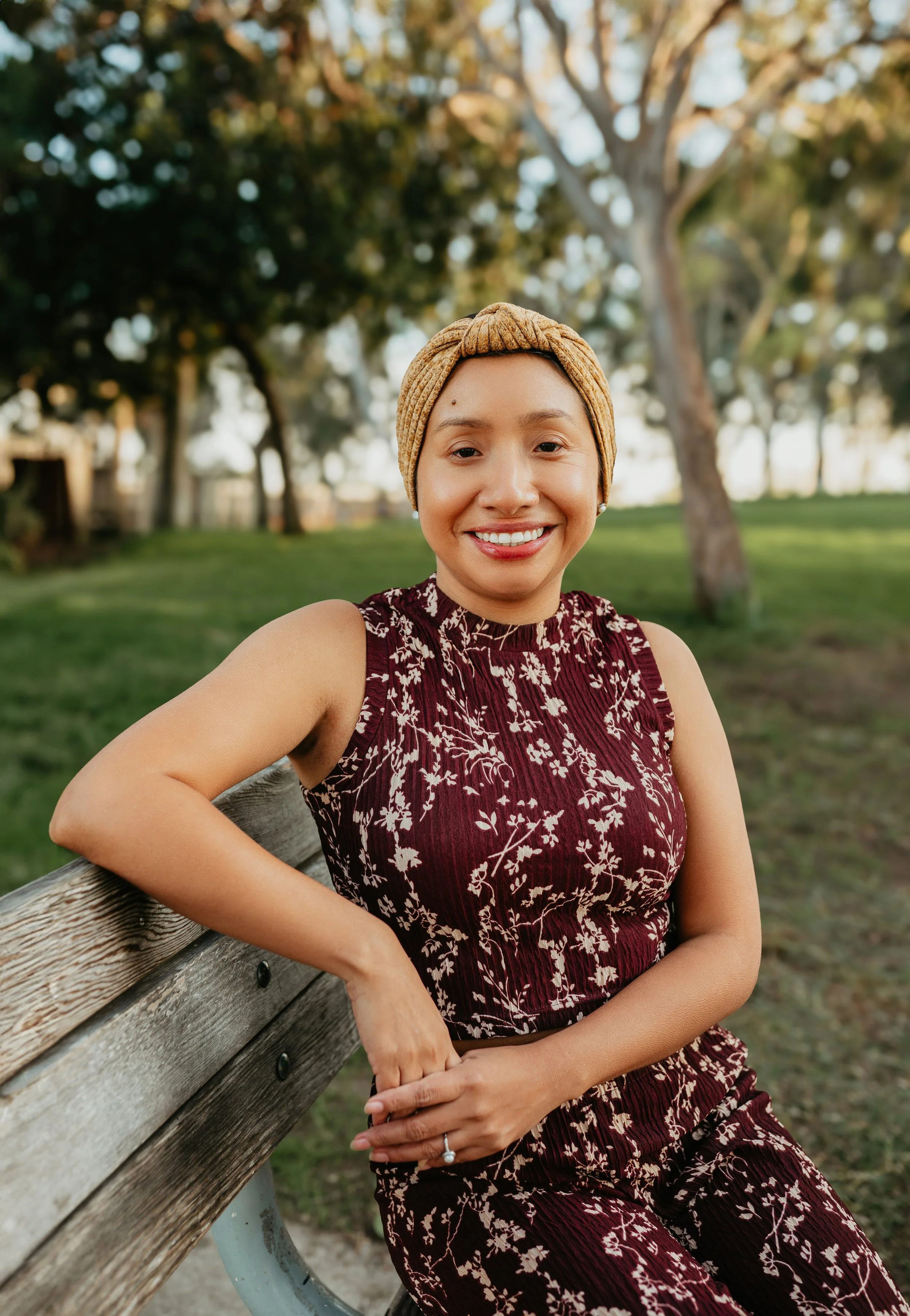 A woman sitting on a park bench, smiling, wearing a patterned burgundy dress and a beige headwrap, with trees and grass in the background.