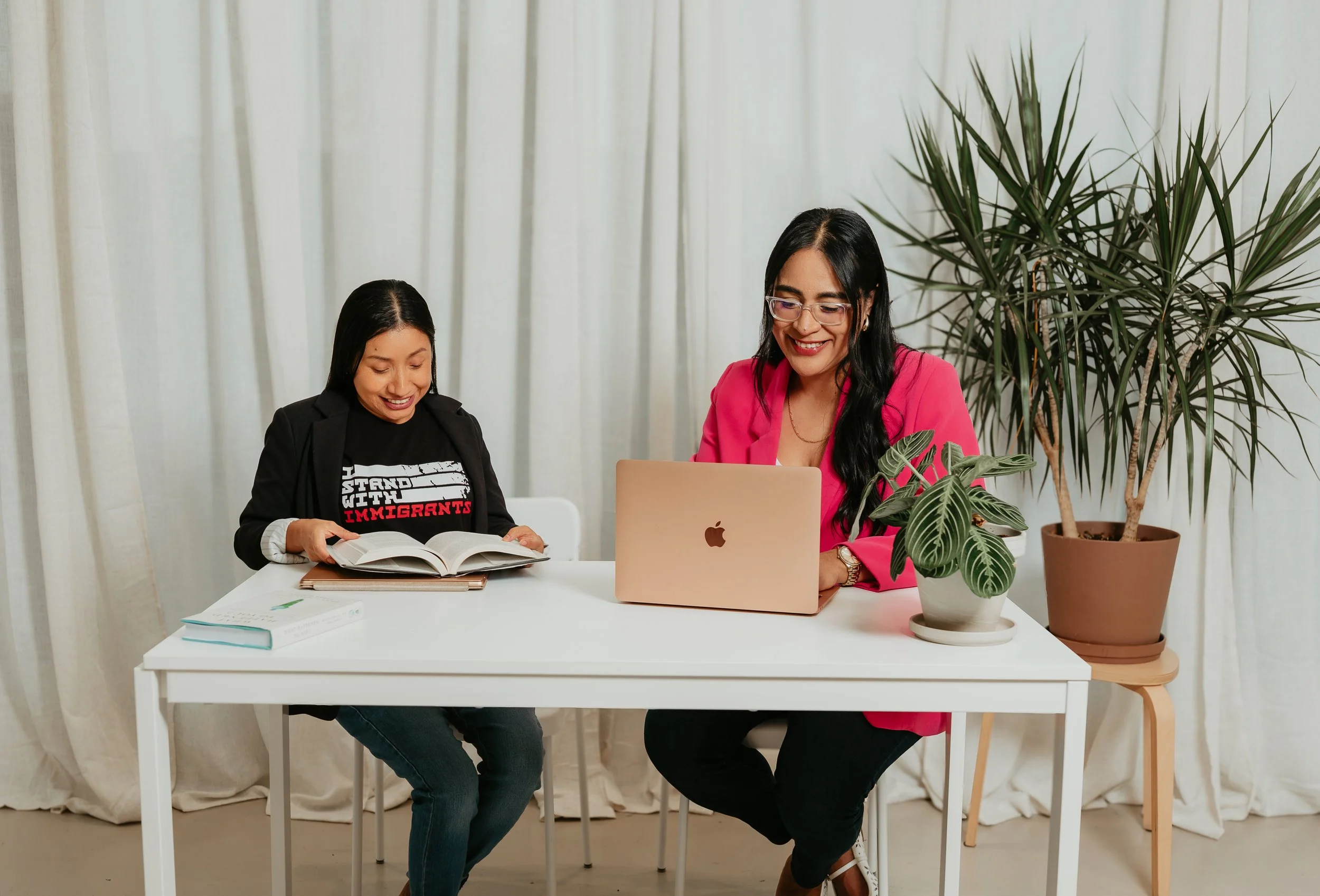 Two women sitting at a white table, one reading a book and the other working on a MacBook, with a large plant in the background.