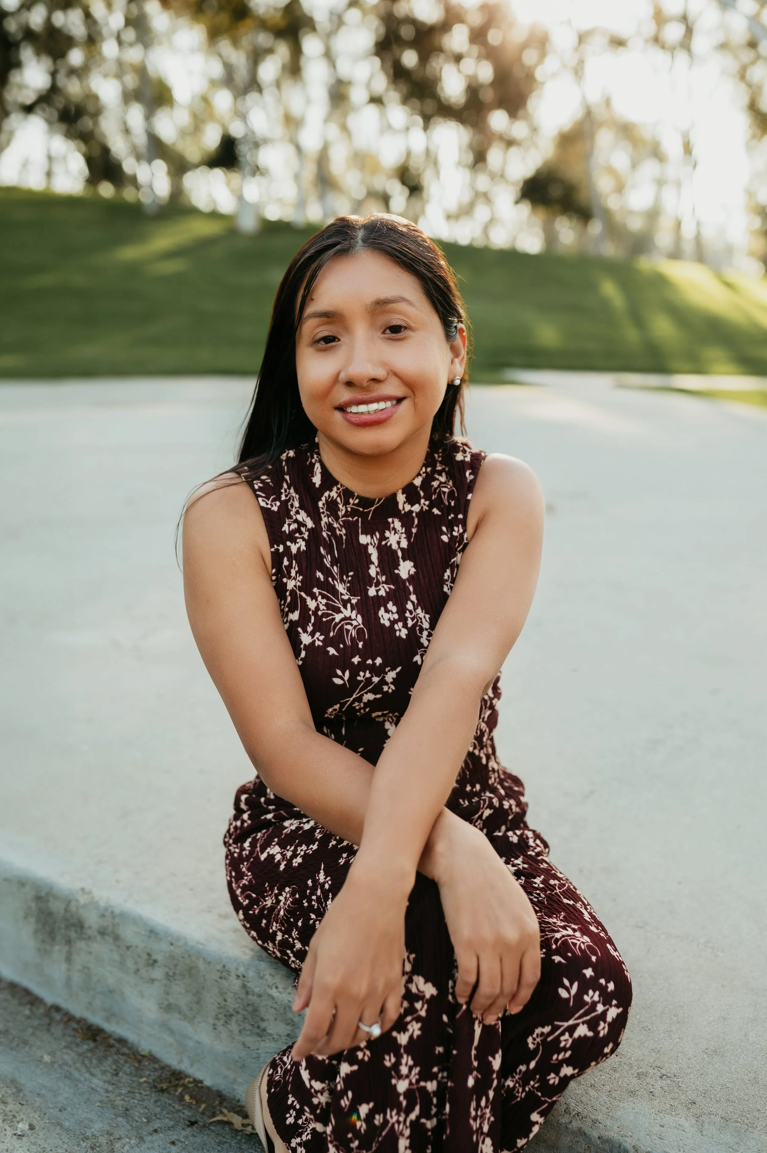 A young woman with long dark hair, wearing a sleeveless dark floral dress, sitting on a curb in a park during sunset, smiling at the camera.