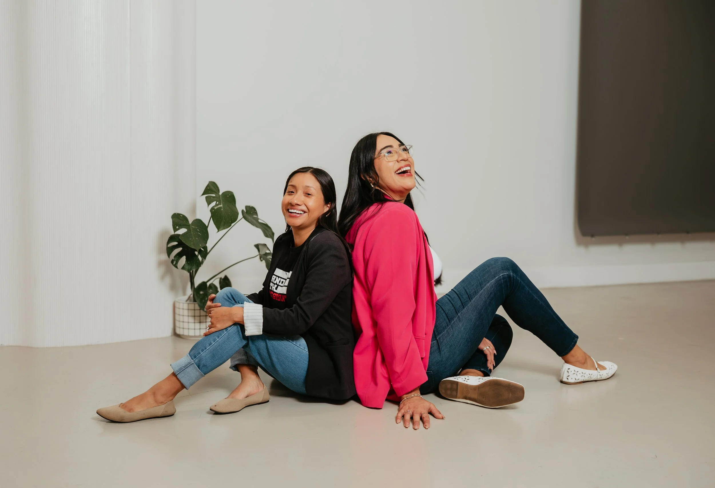 Two women sitting on the floor back to back, smiling and laughing, in a bright room with a large green plant in a pot nearby.