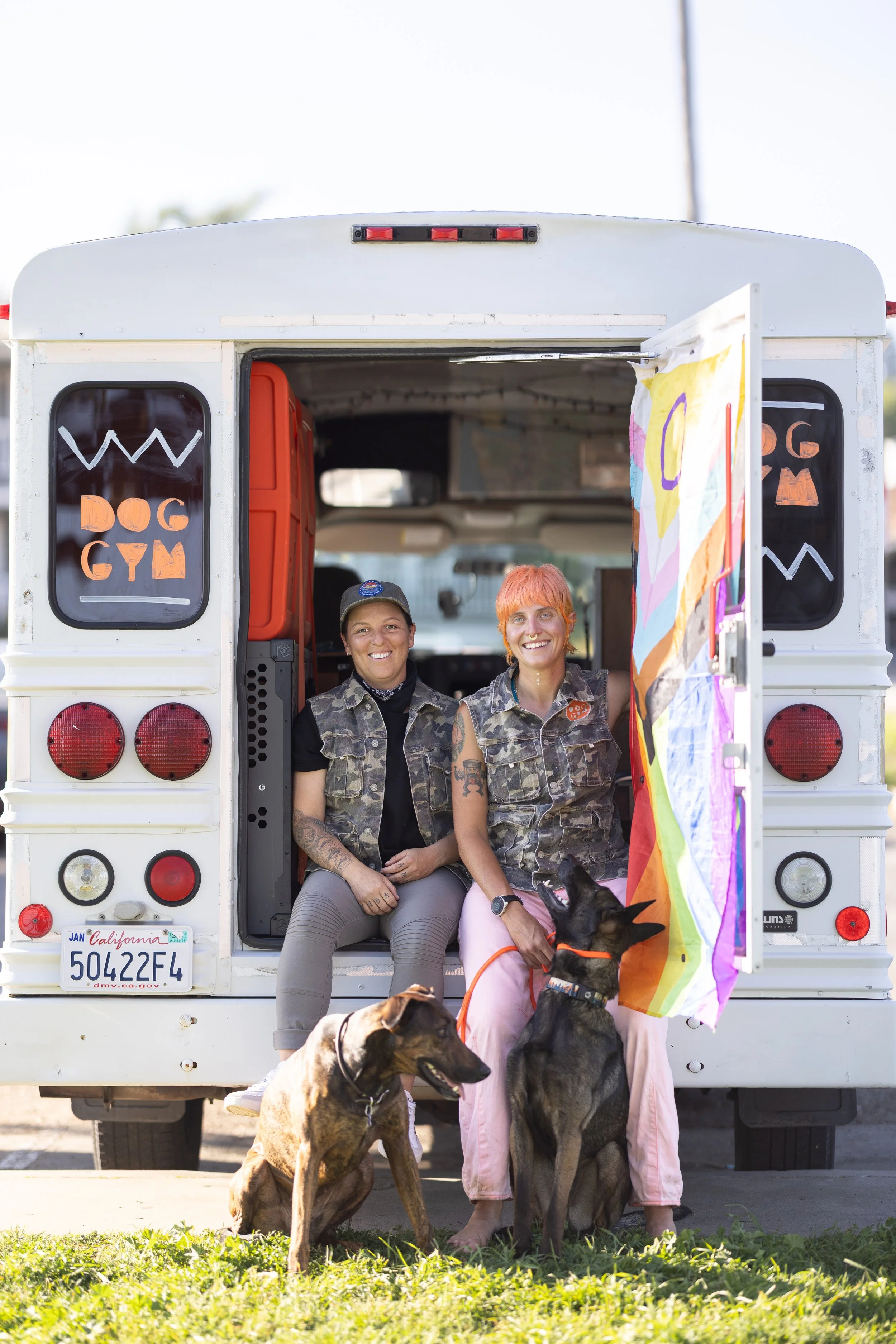 Two people sitting in the back of a white van with a hand-painted sign reading 'Dog Gym,' along with two dogs sitting, one looking up at the person with the rainbow flag, and the other looking to the side. Both people are smiling, wearing casual clot