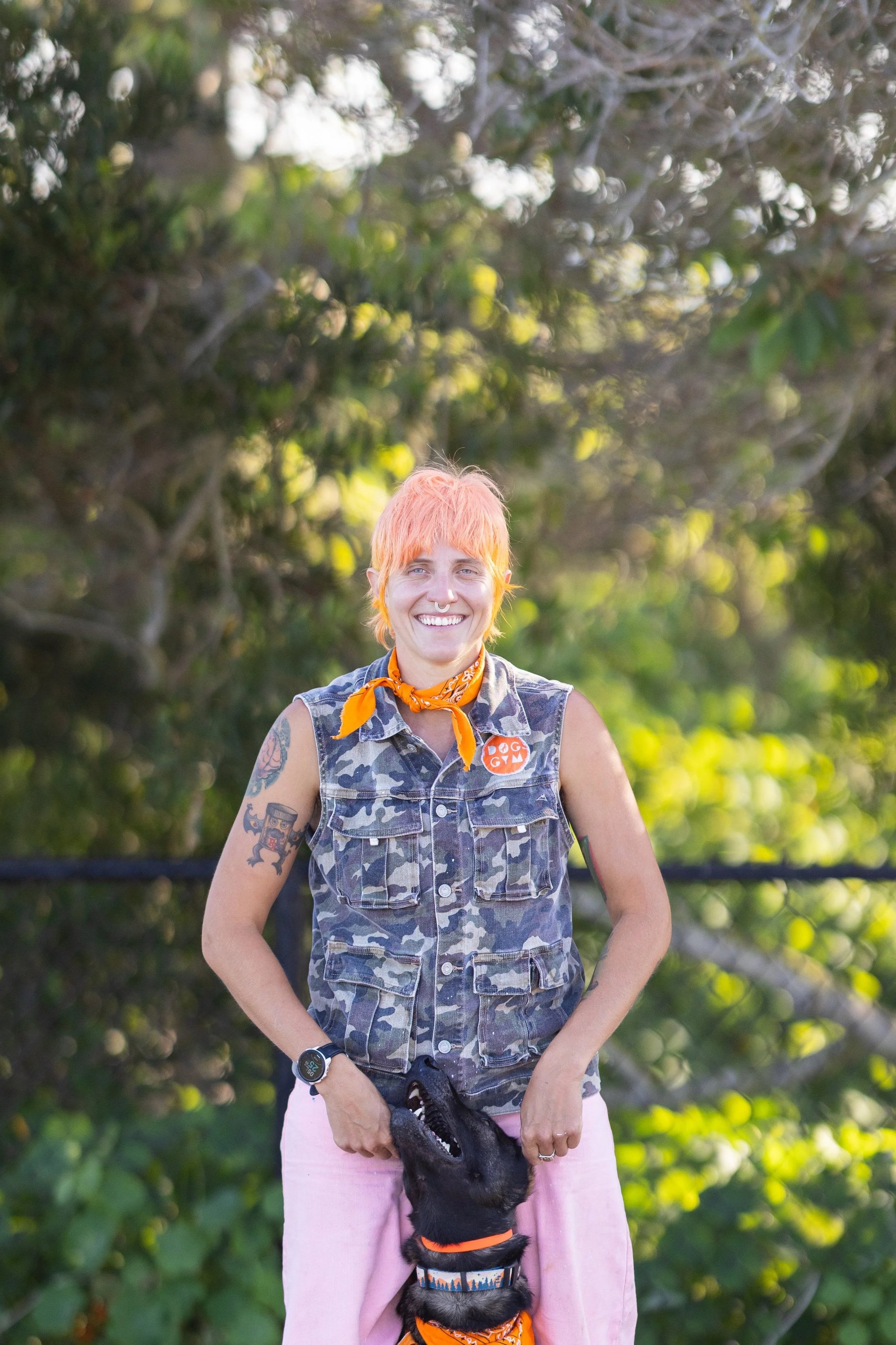 A person with short, pink hair smiling outdoors, wearing a camouflage vest, pink pants, and an orange bandana, holding a black dog wearing an orange collar.