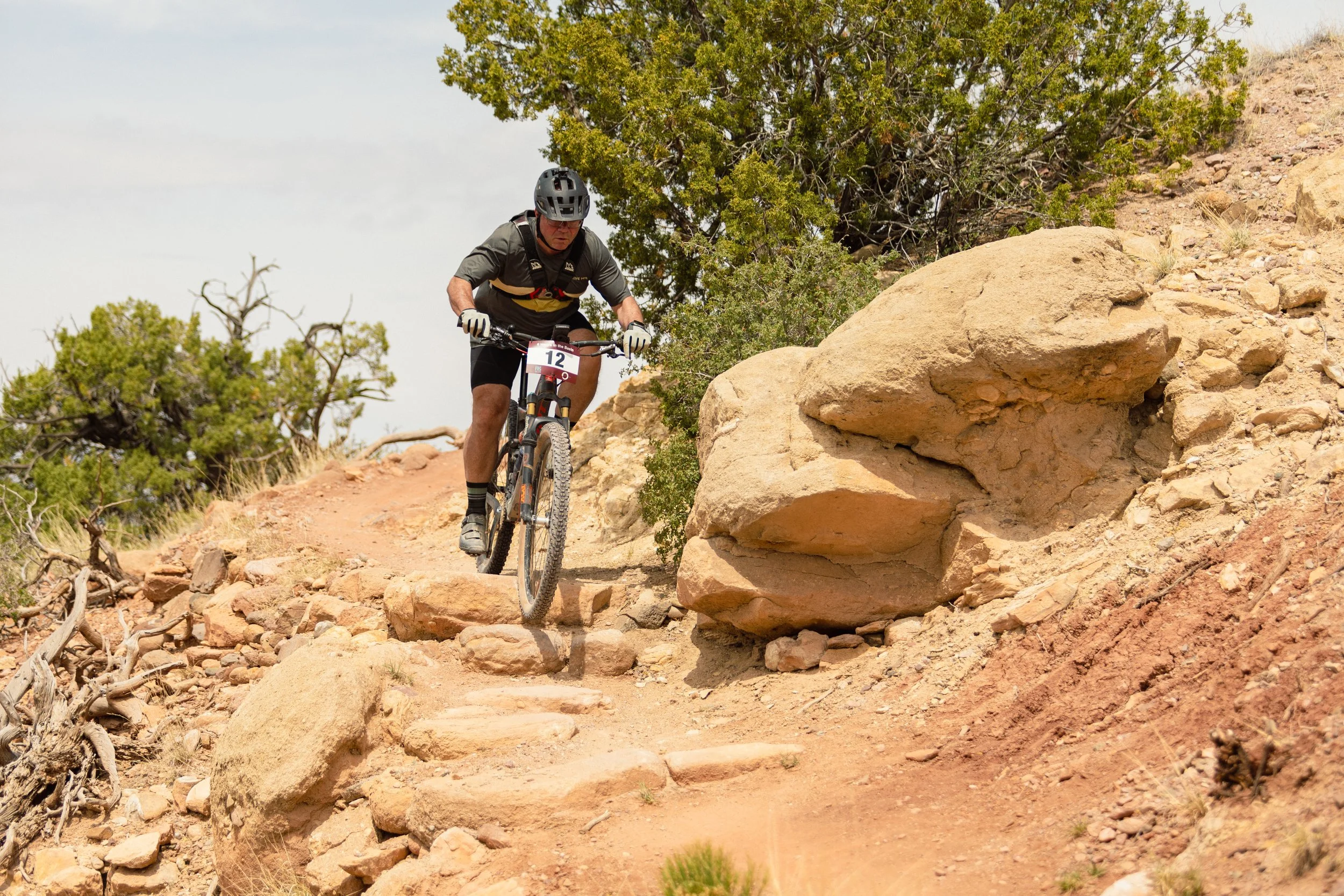 A person riding a mountain bike down a rocky dirt trail with trees and rocks around, wearing a helmet and athletic clothing.