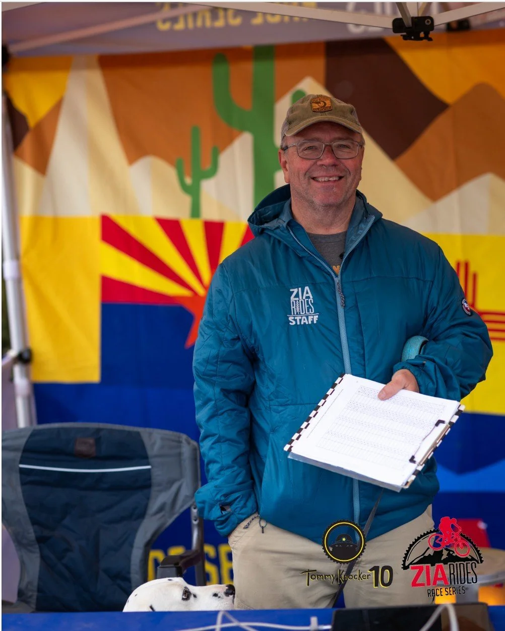 A smiling man wearing glasses, a cap, and a blue jacket with 'Zia Rides Staff' printed on it, holding a clipboard. There is a small dog in a stroller in front of him. The background features a desert scene with cacti and mountains, and logos for 'Tommy Knocker 10' and 'Zia Rides Race Series'.