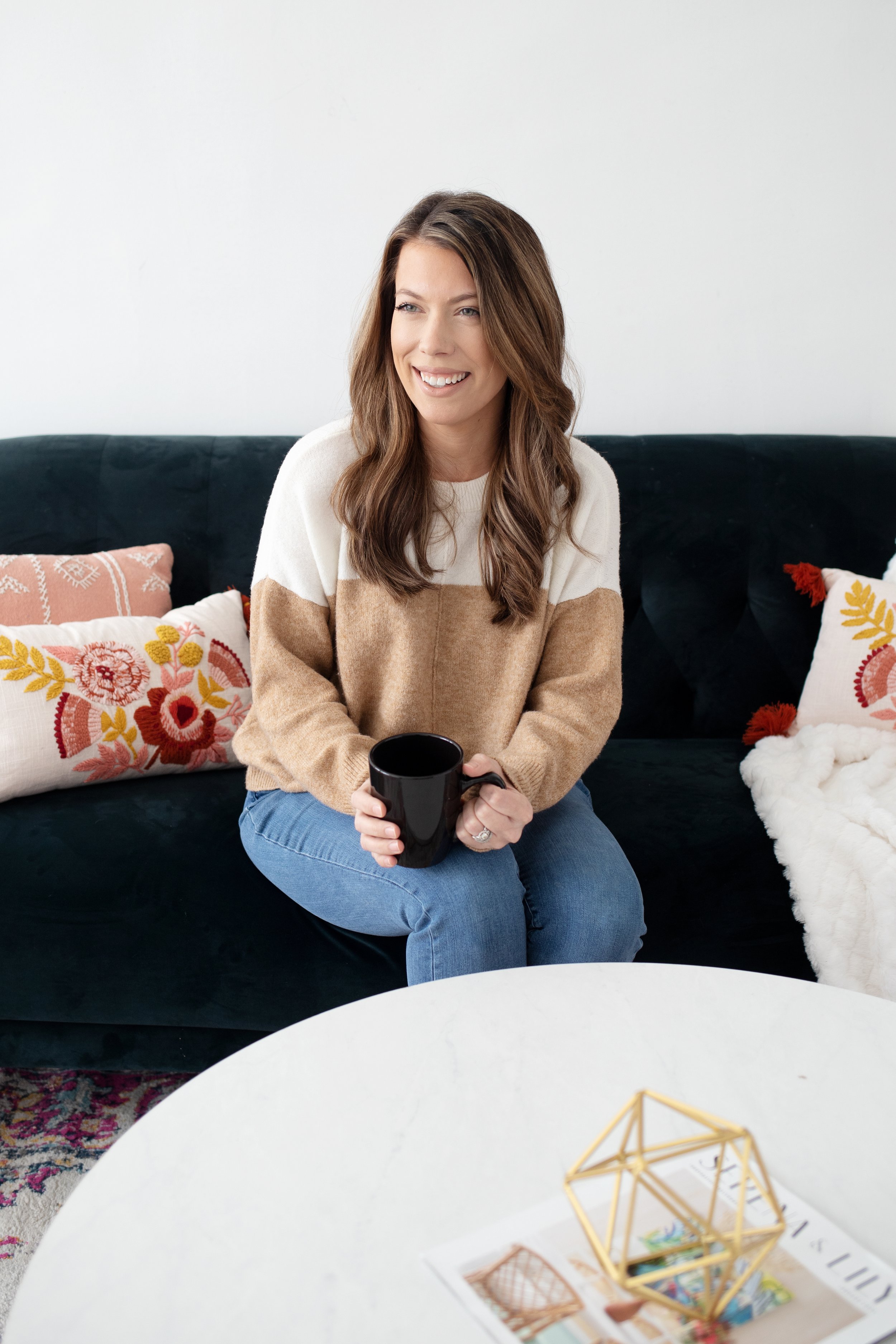 A woman with long brown hair sitting on a black sofa, holding a black mug, smiling, in a cozy room with embroidered pillows and a white table with a decorative gold geometric object.