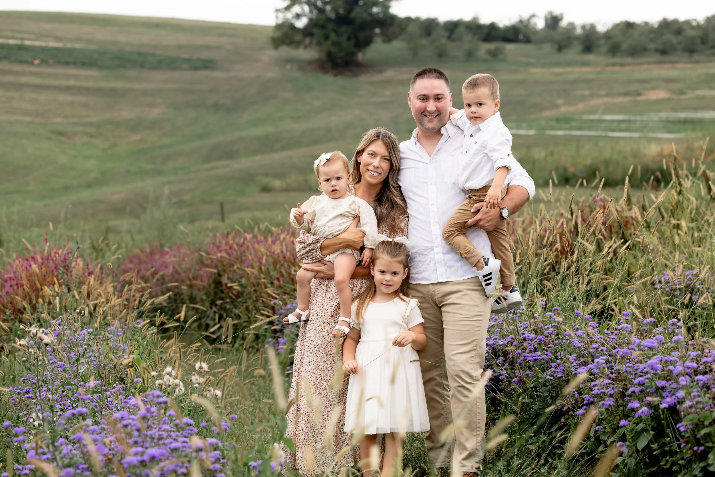 A family of six poses together outdoors in a field of wildflowers and tall grass, with rolling hills in the background.