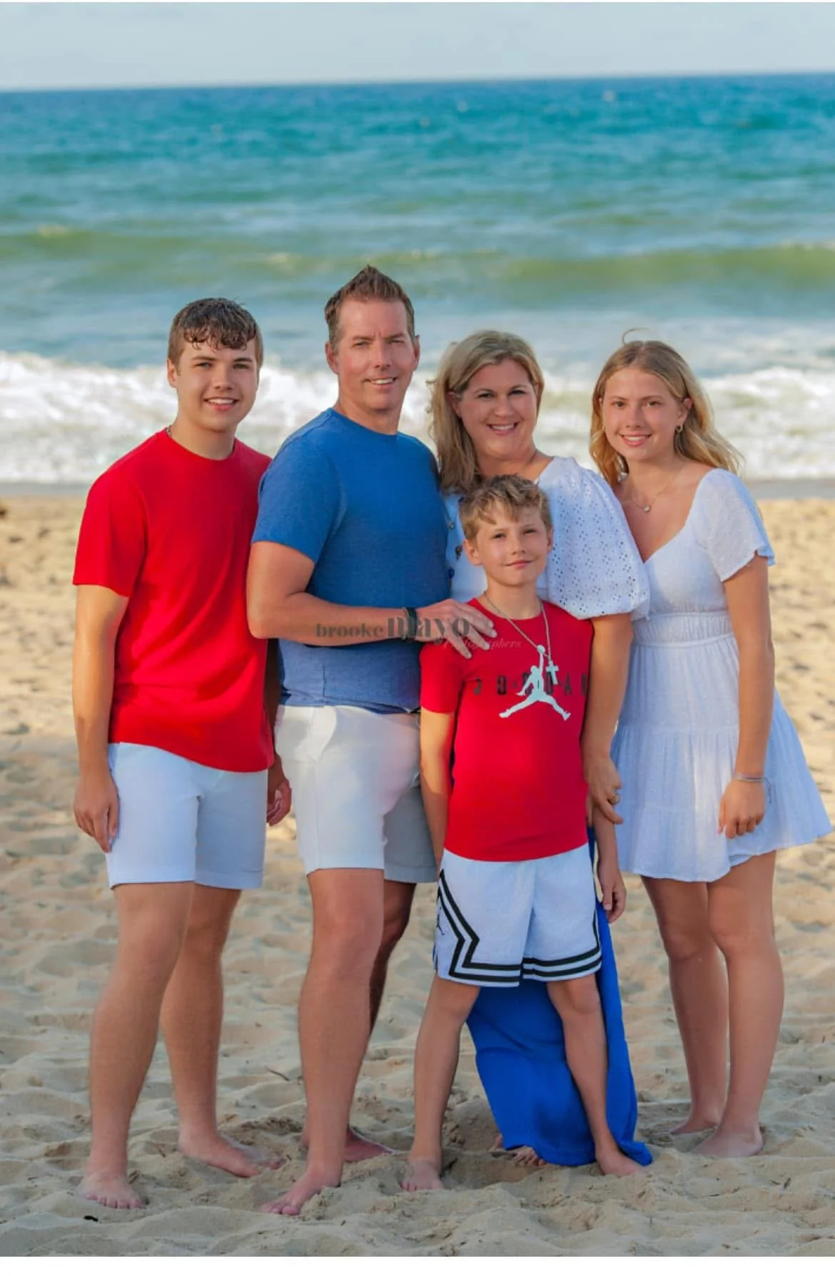 A family of five standing on a sandy beach with the ocean and blue sky in the background, smiling at the camera.