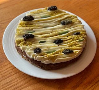 Gâteau rond à la crème, décoré de mûres et de zestes d’agrumes, servi sur une assiette blanche en bois.